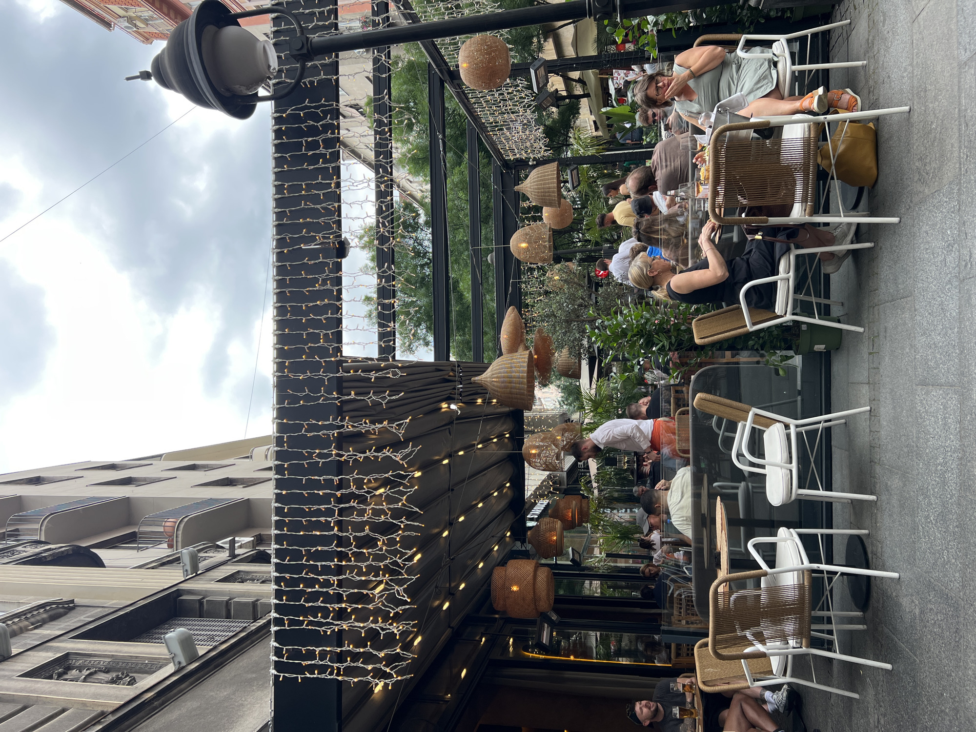 The image depicts an outdoor cafe in Belgrade, Serbia. The cafe has a black metal structure adorned with strings of small lights and woven basket-like lamps hanging from the ceiling. Several people are seated at tables, some with drinks and food. One woman is wearing a black tank top and another is wearing a light green dress. Some are interacting, while others appear to be relaxing. The cafe is situated on a sidewalk next to a building with multiple windows. Above, the sky is cloudy. A street lamp is visible in the foreground. There are different chairs in the cafe, some are white, some are made of wooden material. The location appears to be a well-visited spot, with people occupying several tables. The environment is urban, with the architectural elements of buildings visible in the background. A man sits on the ground on the lower left corner.