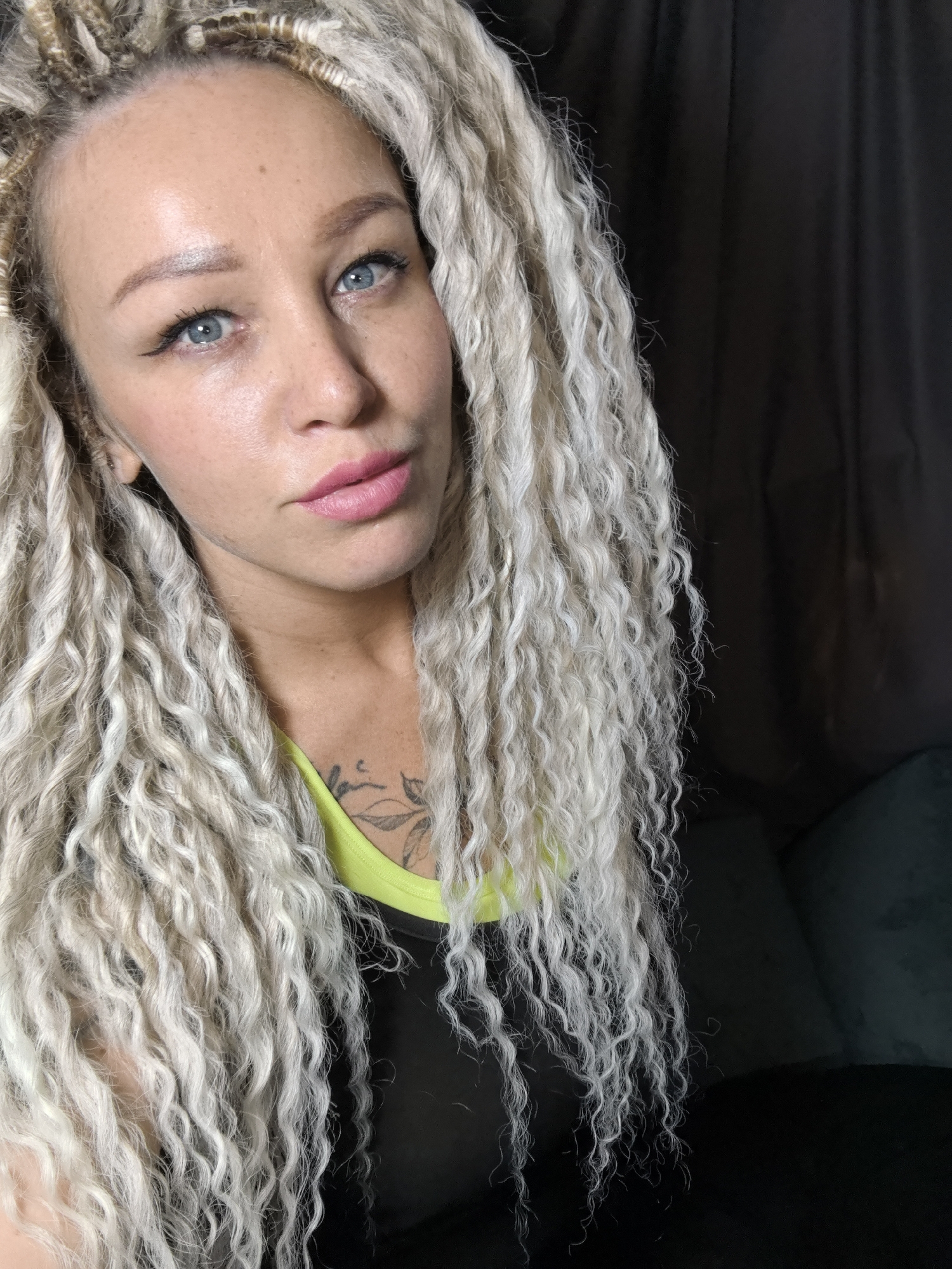 A woman is depicted in a medium close-up, chest-up portrait, looking directly into the camera. She has striking long, very light blonde or white dreadlocks or braided extensions, which are predominantly wavy and cascade over her shoulders and chest. Her bright blue eyes are emphasized with winged eyeliner, and her light brown eyebrows are well-defined. Her fair skin shows a scattering of freckles across her forehead and cheeks, and her lips are painted with pink lipstick.
She is dressed in a black top featuring a neon yellow or green trim around the neckline, suggesting athletic wear. A dark-colored tattoo, appearing to be a floral or leaf design, is visible on her upper chest/shoulder, partially obscured by her hair.
The background is dark and out of focus, indicating an indoor setting. On the right, a dark fabric, possibly a curtain or wall, is visible, and on the lower right, a hint of dark, plush furniture can be discerned. The lighting is soft and even, highlighting her features without harsh shadows. Her expression is direct and composed. There is no visible text in the image. This photograph was taken in Noviy, Russia.