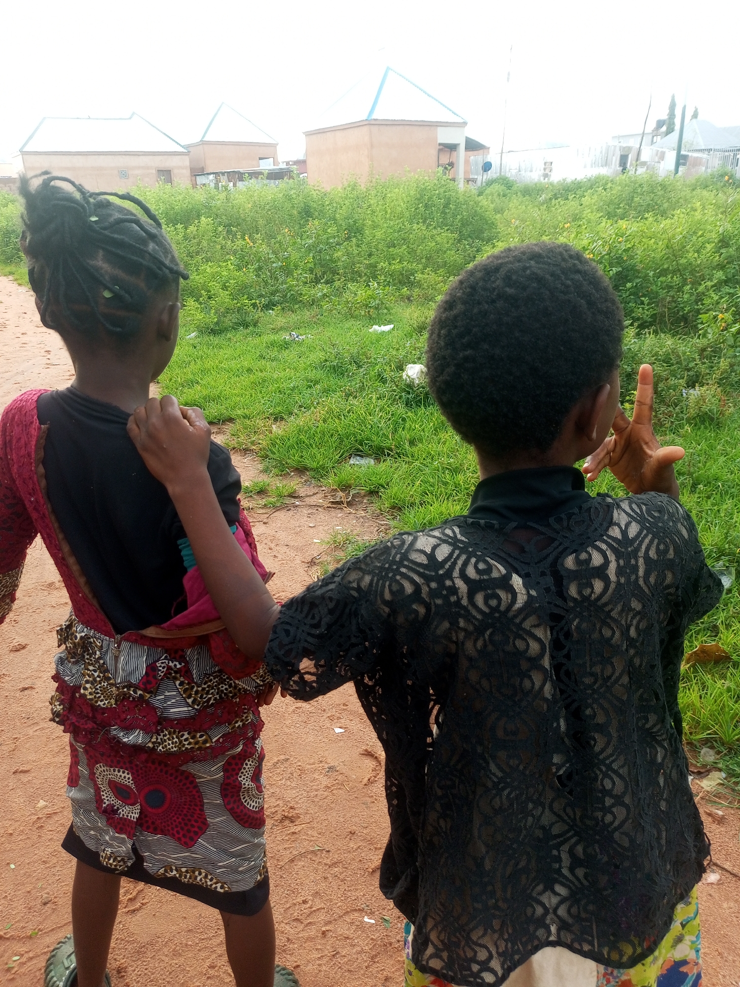 In Tempure, Nigeria, two young children stand facing away from the camera on a dirt path. The child on the left has braided hair and wears a black shirt with a red and patterned fabric skirt. The child on the right has short, curly hair and is wearing a black, lacy top over a patterned skirt. In the background, there is dense green vegetation and several buildings with light-colored walls and light-colored roofs. The sky is overcast.