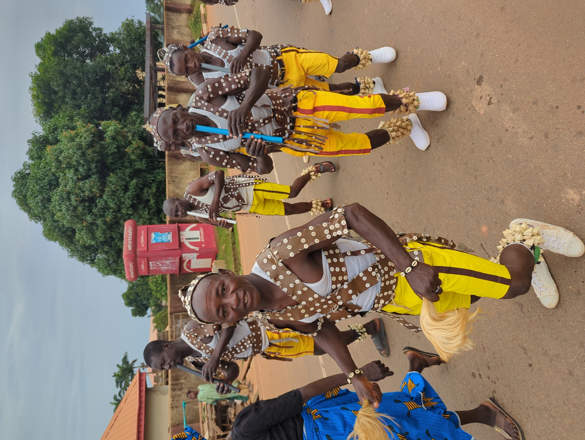 A group of men are seen in Kafanchan B, Nigeria, seemingly performing a traditional dance. The men are dressed in matching outfits consisting of yellow pants with brown stripes, white tank tops, and brown buttoned vests. They are wearing beaded headbands and bracelets, and some have ankle decorations. Several of the men are playing long, blue flutes. The men are standing on an asphalt road with buildings and trees in the background. A red "smartcash" ATM machine is visible in the background. The weather appears overcast.