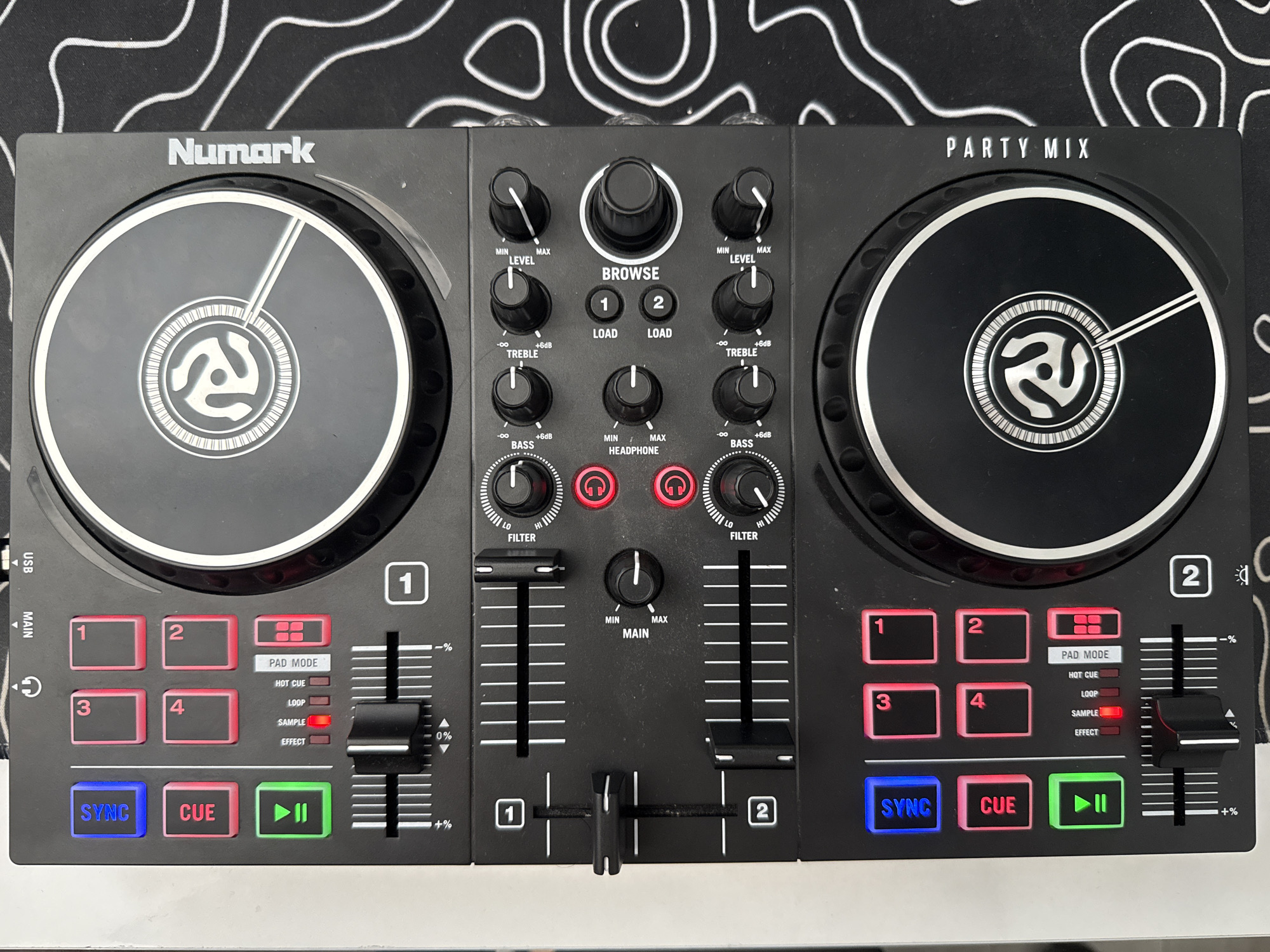 The image shows a black Numark Party Mix DJ controller placed on a black mat with white topographic line art. The DJ controller is positioned horizontally and occupies the majority of the frame. It features two circular jog wheels, each with a stylized circular design in the center. Above the left jog wheel are several illuminated buttons: a blue "SYNC" button, a red "CUE" button, and a green button with a play/pause icon. To the right of these are four red square buttons labeled "3", "4", "1", and "2". Below the "CUE" button are smaller indicator lights labeled "EFFECT", "SAMPLE", and "LOOP", with the "SAMPLE" light illuminated red. There are also several sliders and knobs for controlling audio. Above the right jog wheel, the text "Numark" is printed vertically. Below the right jog wheel, the text "PARTY MIX" is printed vertically. The environment appears to be indoors, with the DJ controller set on a flat surface. There is no indication of the city of Burgas, Bulgaria in the image itself.