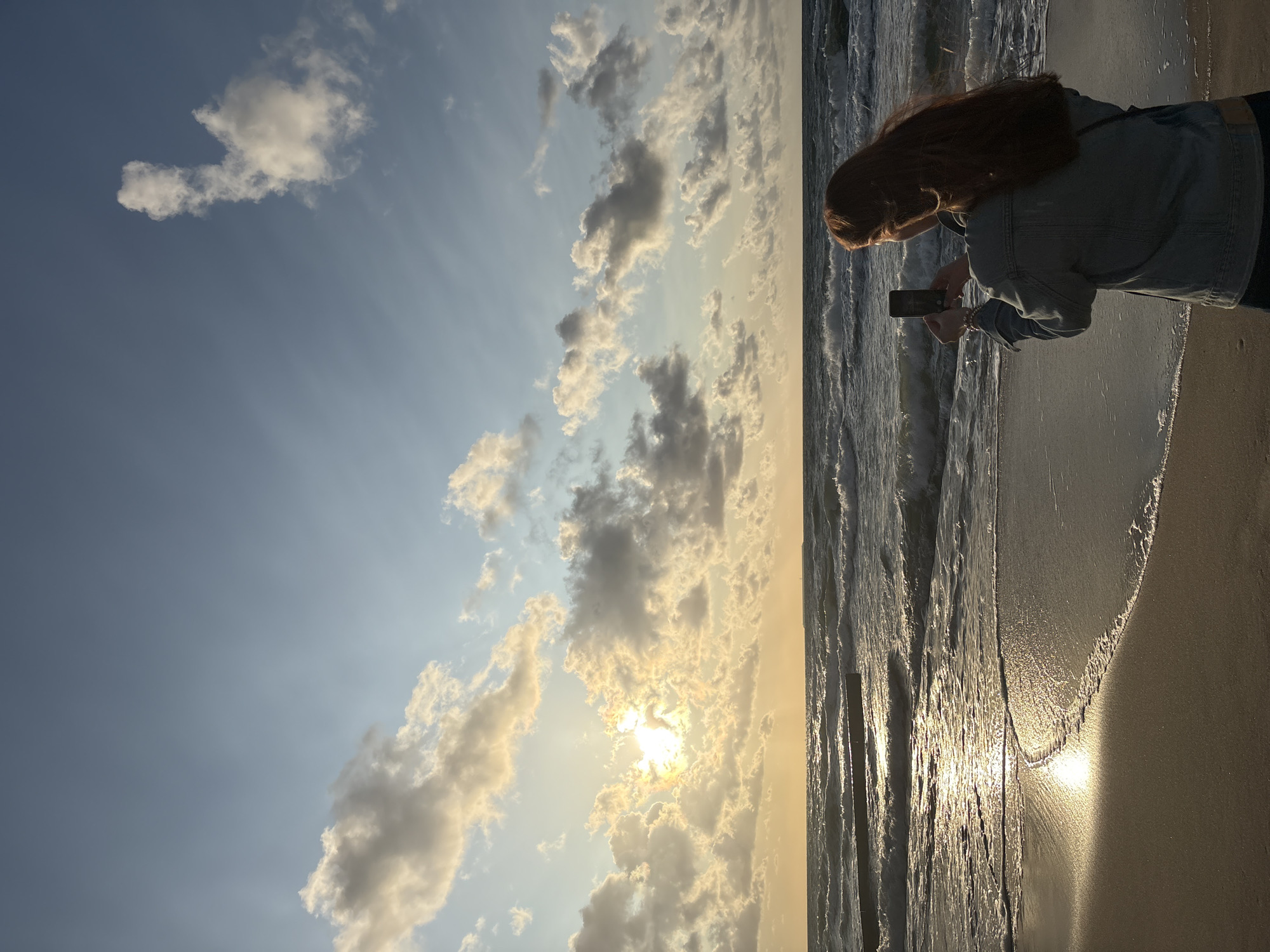 An overhead shot shows a woman with long, auburn hair, wearing a denim jacket, standing on a sandy beach and taking a photograph with her cell phone. The woman is facing the ocean, with her back to the camera. She has a bracelet on her left wrist. The ocean waves are rough, with white foam on top. The sun is setting behind a cluster of clouds on the left side of the image, casting a golden glow on the water and the clouds. The sky is blue with scattered clouds. In the distance, there are structures extending into the ocean, possibly groins. The location is Zelenogradsk, Russia.