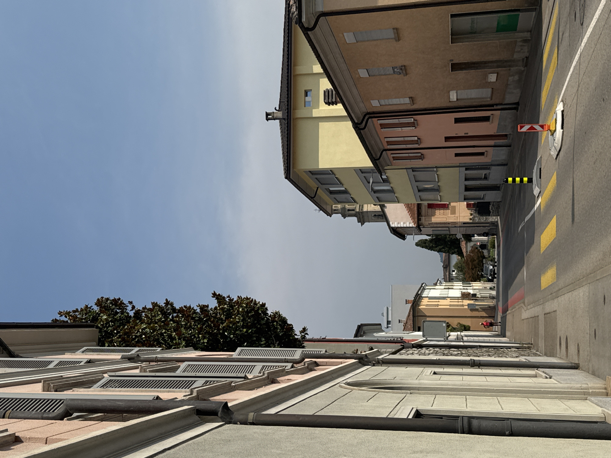 The image depicts a street scene in Balerna, Switzerland, under a clear blue sky. A narrow street is lined with buildings on both sides. On the left, a building with a pink facade and gray shutters is prominent, with a large tree with dark green foliage extending from behind it. The building on the right has facades in yellow, pink, and tan colors, with multiple windows and doors visible. The street is paved with asphalt and features yellow crosswalk markings. Several traffic cones and bollards are placed on the road, some with reflective orange and white markings. In the distance, a car is seen driving down the street. A lone figure, appearing to be a woman in athletic wear, is walking on the sidewalk on the left side of the street. Further down the street, another building with a distinctive bell tower is visible. The overall impression is a quiet, urban environment on a sunny day.