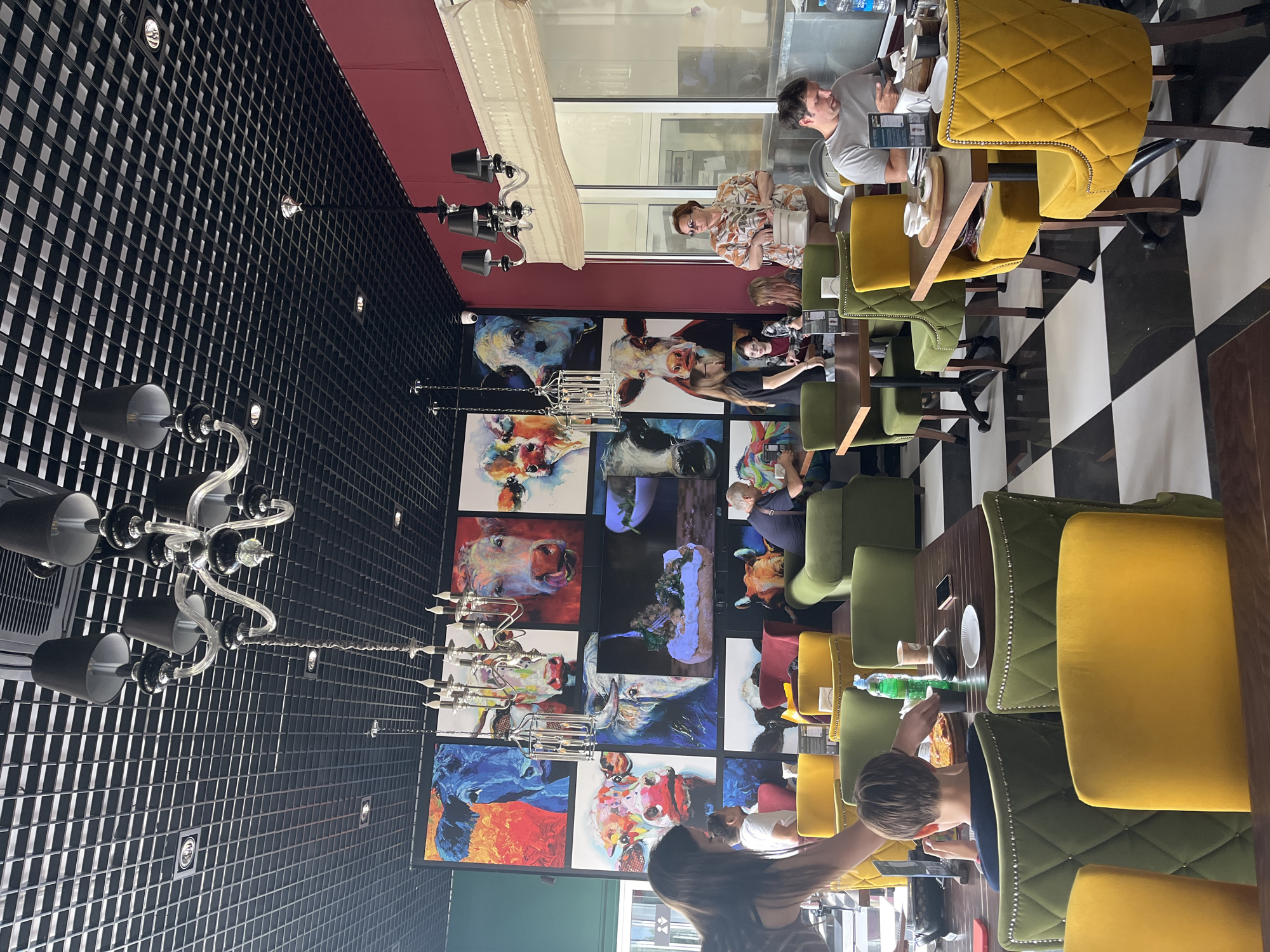 The image depicts the interior of a restaurant, likely located in Akhlebaikha (Kovriginskii S/S), Russia, characterized by its modern and artistic decor. The dominant feature of the ceiling is a black grid pattern with recessed lighting and two ornate chandeliers. The walls are adorned with a vibrant collage of large paintings, predominantly featuring stylized portraits of cows in various colors and expressive poses.
The restaurant's seating arrangement includes tables with dark wood tops and chairs upholstered in bright yellow and deep green velvet, many with decorative nailhead trim. The floor is a striking black and white checkerboard pattern.
Several individuals are visible within the restaurant. In the foreground, a young person with short brown hair is seated at a table, looking at their phone while a pizza sits before them. To their left, a woman with long dark hair, seen from the back, appears to be interacting with the young person or the table. In the middle ground, a group of people are seated at a table, including a man in a grey t-shirt looking at his phone, and a woman with sunglasses and a floral-patterned shirt holding a purse. Further back, other patrons are seated at tables and conversing. The overall atmosphere suggests a casual dining experience.