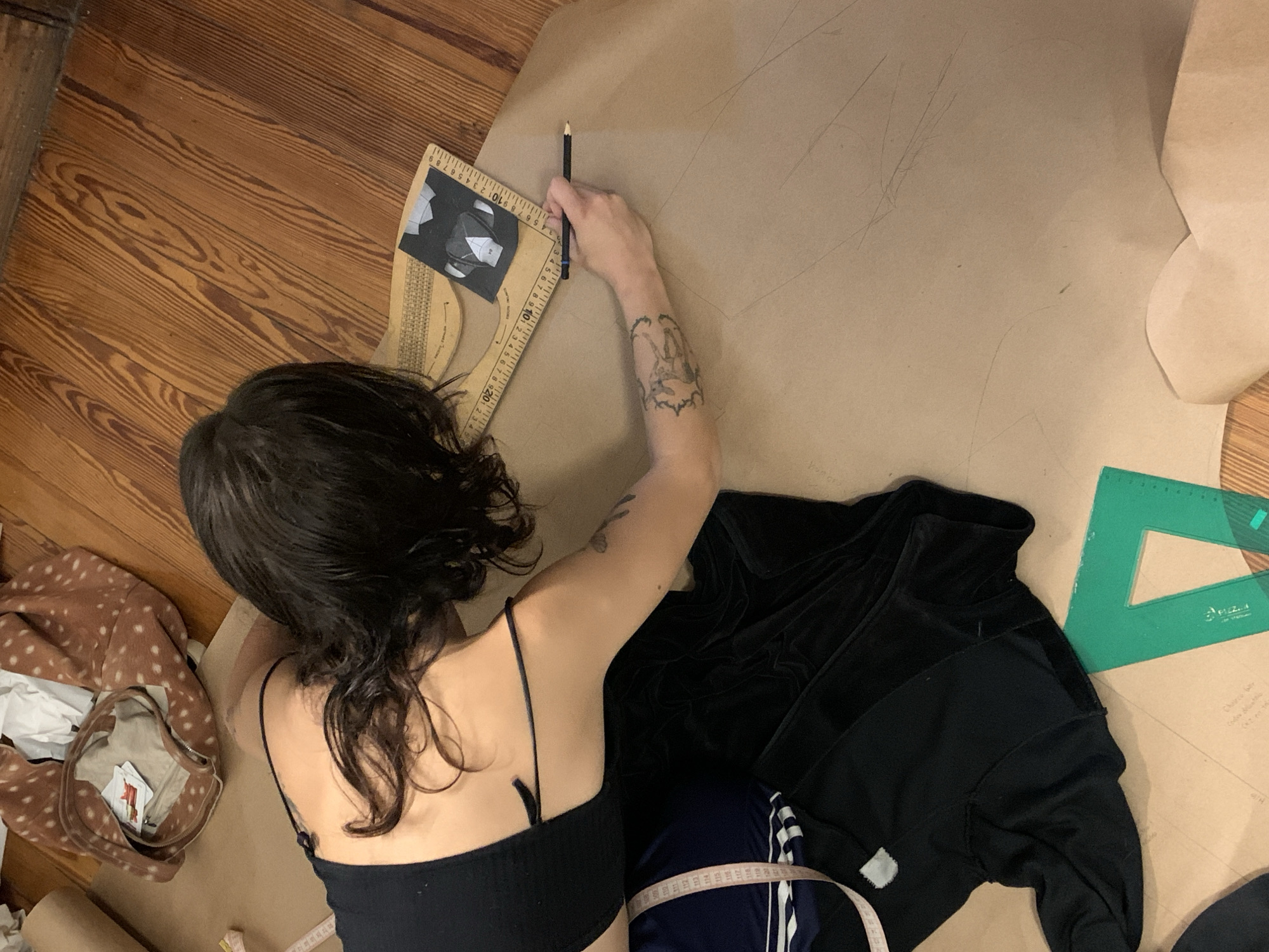An overhead shot captures an individual engaged in pattern drafting on a wooden floor. The person, seen from behind and slightly to the side, has dark, wavy hair and is wearing a black spaghetti strap top. Their right arm, which bears a tattoo featuring figures within a band, holds a pencil and is actively marking lines on a large sheet of brown kraft paper spread across the floor.
A large wooden pattern-making ruler, marked with measurements and "MADE IN U.S.A.", is positioned to their left, assisting in the precise drawing. A small black rectangular object, possibly a reference pattern piece displaying numbers "54" and "44", rests on this ruler. Another green plastic set square, with the name "PIZZI" visible, lies on the brown paper to the right.
Various garments and materials surround the workspace. A black jacket, possibly made of velvet or velour, is located to the right of the person. Beneath it, a blue and white striped garment (suggesting athletic wear) is partially visible, draped with a fabric tape measure. To the person's left, a brown, fawn-spotted garment is crumpled alongside some white paper and a roll of brown paper.
The setting is indoors, likely a creative studio or home, characterized by warm-toned wooden floorboards. The activity suggests a focused crafting endeavor, such as fashion design or sewing. No specific time of day or emotional state is discernible beyond the individual's concentration. While the provided context indicates Buenos Aires, Argentina, no direct visual cues in the image confirm this location. Faint text "Yor wore..." can be seen on the brown paper near the bottom left.