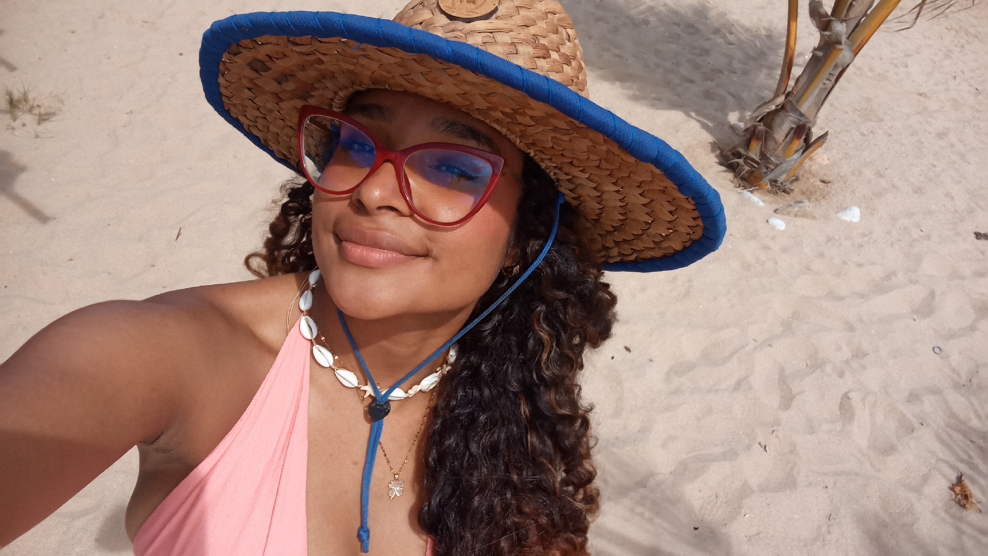 A young woman with curly brown hair wears a straw cowboy hat with a blue band and red-framed glasses. She is dressed in a pink swimsuit and a seashell necklace. She smiles as she takes a selfie on a sandy beach. In the background, a small palm tree trunk emerges from the sand. The scene appears to be outdoors on a sunny day.
