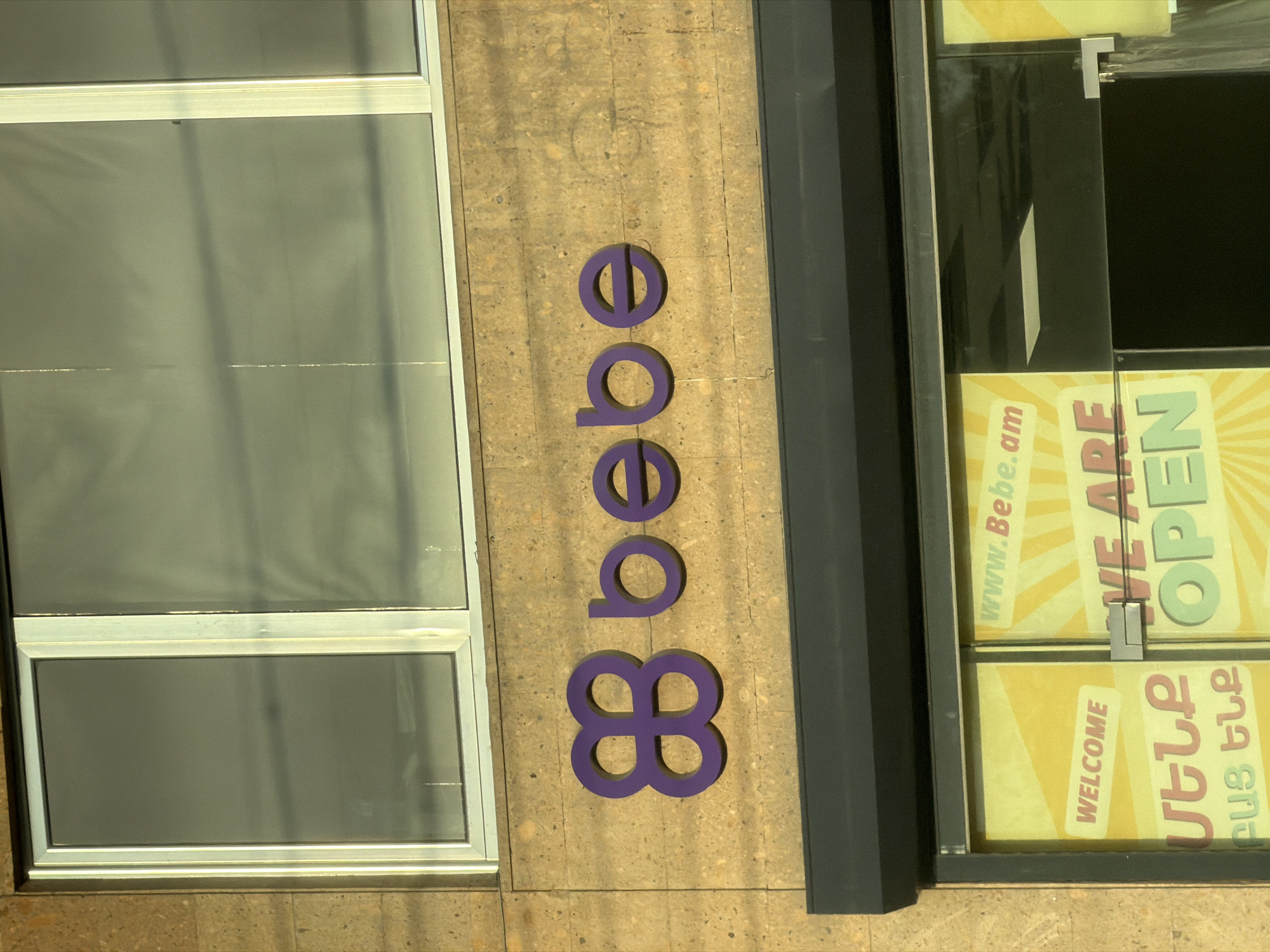 This media file shows the exterior of a Bebe store in Yerevan, Armenia. The main subject is the Bebe logo, rendered in large, purple, three-dimensional letters vertically along a stone-clad wall. The letters spell "bebe" with the "b" appearing twice, followed by two circles and a four-leaf clover-like symbol.
To the left of the logo is a large window with a silver frame, divided into two sections. The top section appears to be darker, possibly tinted or covered, while the bottom section is clearer, revealing a muted reflection of the environment.
To the right of the logo, a glass storefront displays advertisements. One sign, visible through the glass, has yellow rays emanating from a central point and features text in green and purple. Partial text visible on this sign includes "WE ARE OPEN". Below this, another sign is visible with yellow rays and text that includes "WELCOME" in white lettering on a diagonal red banner. Below the "WELCOME" sign, there are two lines of text in Armenian script.
The scene is set in daylight, with shadows cast by the architectural elements, suggesting it is a bright day. The stone wall has a textured, speckled appearance. The overall impression is of a modern retail establishment in an urban setting.