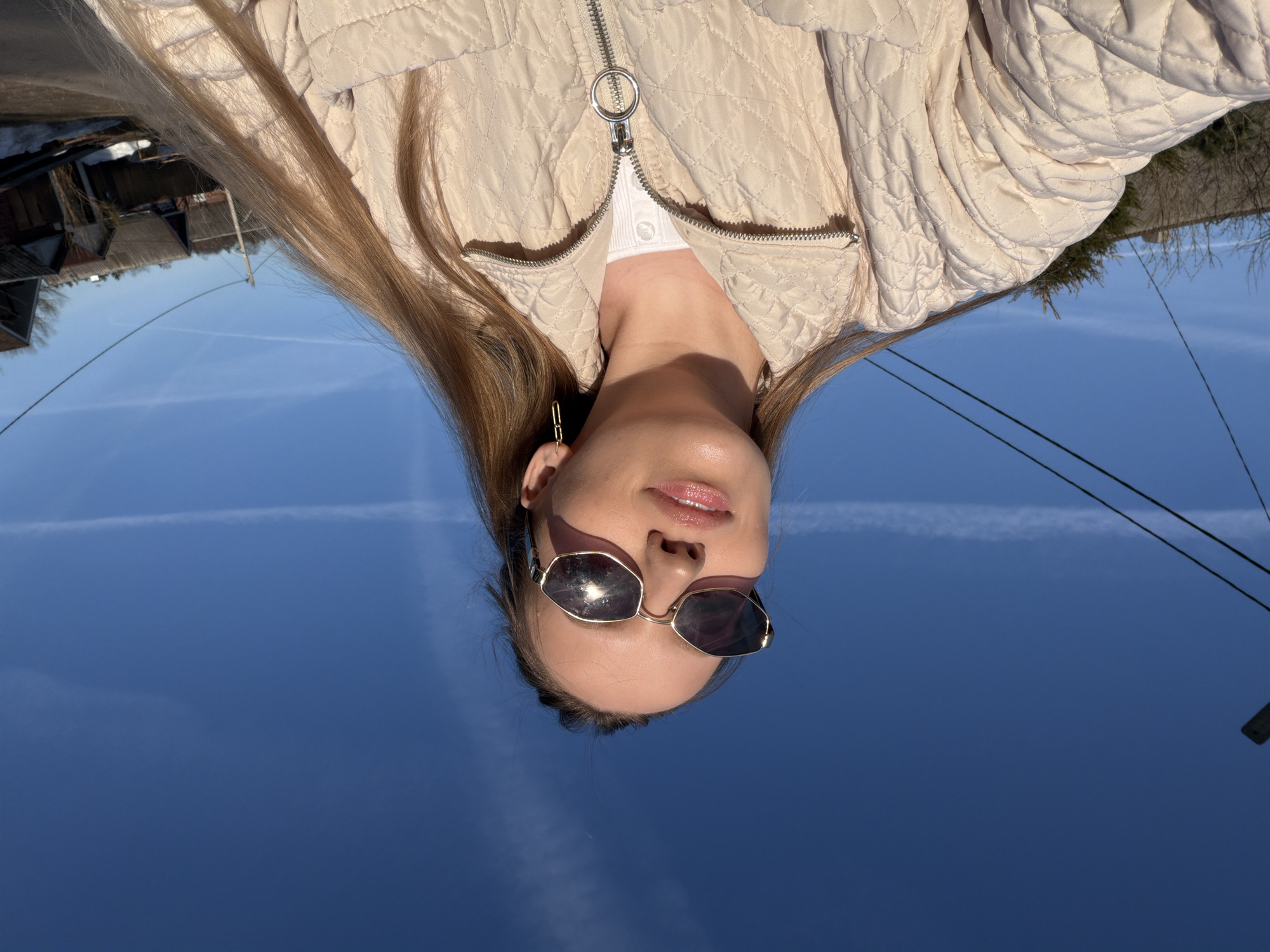 A young woman is pictured from a low angle, facing the camera and wearing sunglasses, a white top, and a quilted beige jacket. She has long blonde hair. The background is a bright blue sky with contrails. Above the sky are buildings, trees and power lines. The location is outdoors, likely in Reshetnikovo, Russia.
