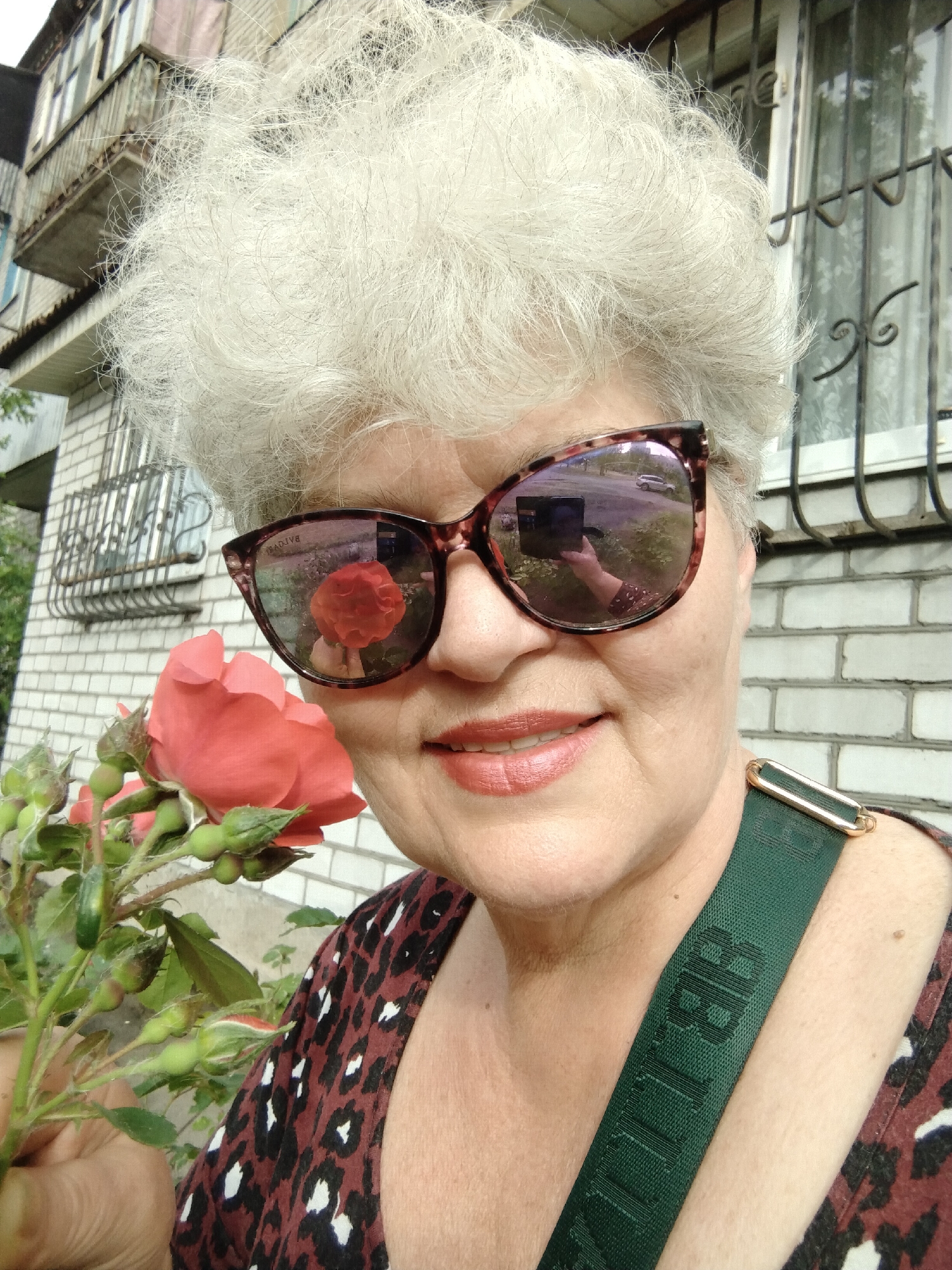 A close-up outdoor selfie prominently features an older woman with striking, voluminous white hair. She is seen smiling gently, her lips adorned with a soft red lipstick, while wearing large brown tortoiseshell sunglasses. She holds a cluster of vibrant pink-red roses and green buds, with one large rose positioned near her left cheek.
The sunglasses offer detailed reflections: the left lens mirrors a red rose from the bouquet, while the right lens reflects an outdoor scene with a road, a distant white car, and her own hand holding a smartphone or camera used to capture the image. She is wearing a dark top with a light animal print pattern. A dark green strap with gold hardware is visible over her right shoulder, bearing the vertically oriented text "BILLIE" (partially obscured). The word "BOLIVE" (reversed) is visible on the upper frame of the left sunglass lens.
The background consists of a light-colored brick building with windows and decorative wrought-iron grilles, typical of a residential area. To the upper left, parts of another building with balconies are visible, providing further context of an urban or suburban environment. The scene is brightly lit, indicating a daytime setting, and the woman's expression suggests contentment or happiness. The location context is Horishni Plavni, Ukraine.
