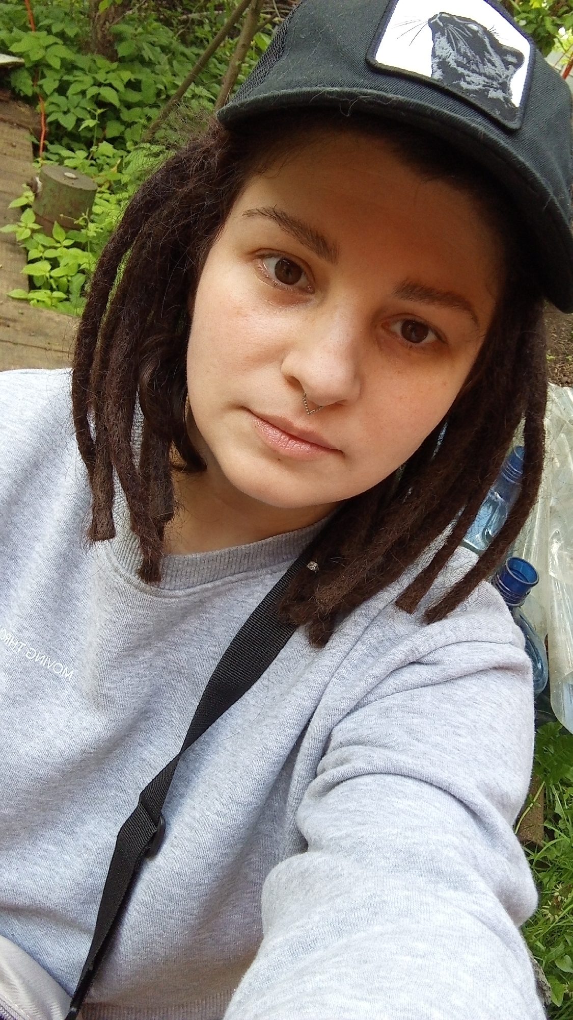 A close-up, slightly angled shot features a person with brown eyes, a nose piercing, and brown dreadlocks wearing a grey sweatshirt with a black strap across their chest. They are also wearing a black cap with a white patch displaying a black cat. The background includes greenery, a wooden structure, plastic bottles, and other indeterminate objects. It's not possible to confirm the location in Юдино, Russia based solely on the provided image.
