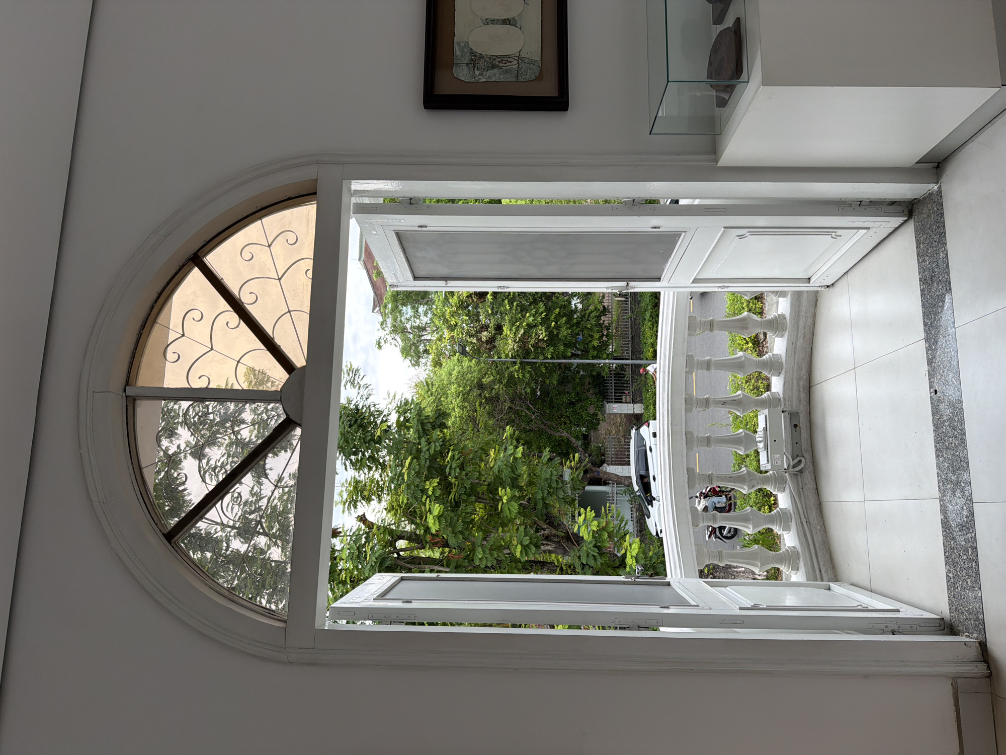 The image captures an interior view looking through an arched doorway with two open doors onto a balcony and a street scene in Thuận Hóa Ward, Vietnam. The doorway features decorative stained glass at the top. The balcony has a white balustrade, and a white electronic device is mounted on it. Beyond the balcony, a street is visible with trees, a white car, a motorbike with a rider, and a person in a red shirt walking. The interior wall is white, and a framed artwork and a glass display case containing artifacts are present on the right side. The flooring inside is tiled.