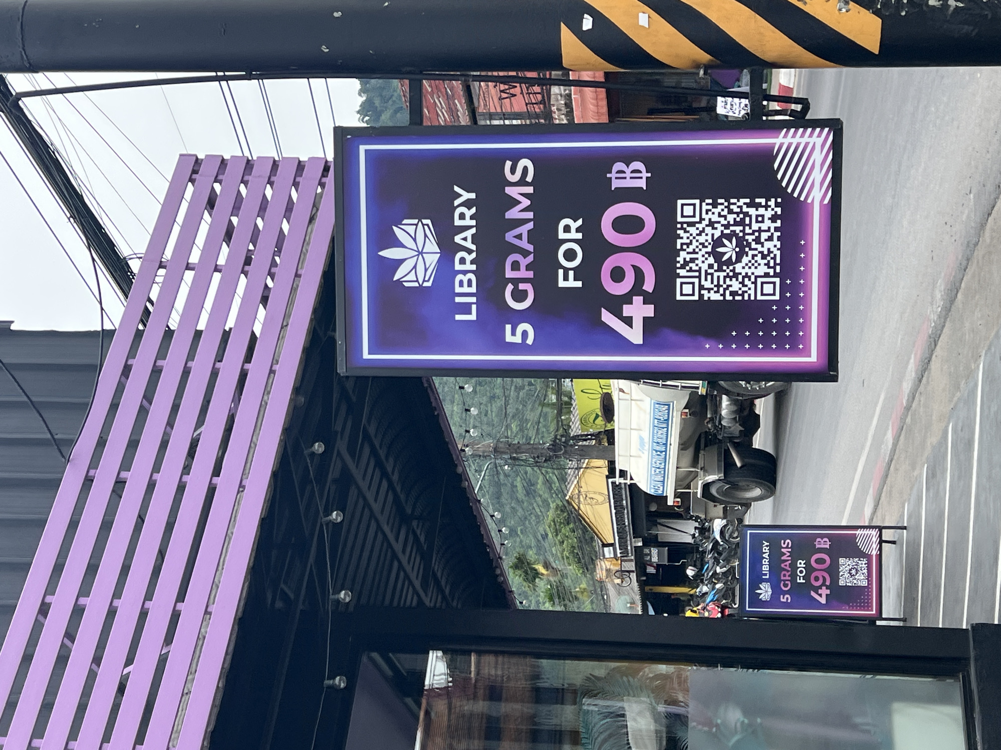 This is a medium-shot, eye-level outdoor photograph taken on a cloudy day on a street in Baan Hua Thanon, Thailand. The foreground features a black pole with yellow and black diagonal stripes on the right side and a dark-colored building on the left. Hanging from a purple structure above the building is a sign that reads "LIBRARY 5 GRAMS FOR 490฿" with a QR code featuring a cannabis leaf. To the left, a smaller, A-frame sign on the sidewalk displays the same information: "LIBRARY 5 GRAMS FOR 490฿". In the background, a white water truck with the words "CLEAN WATER SERVICE" and a phone number is visible on the street. Beyond the truck, there are several buildings with yellow and red roofs, and lush green mountains rise in the distance. A few motorcycles are parked on the left side of the street. The overall scene depicts a commercial area, likely a dispensary.