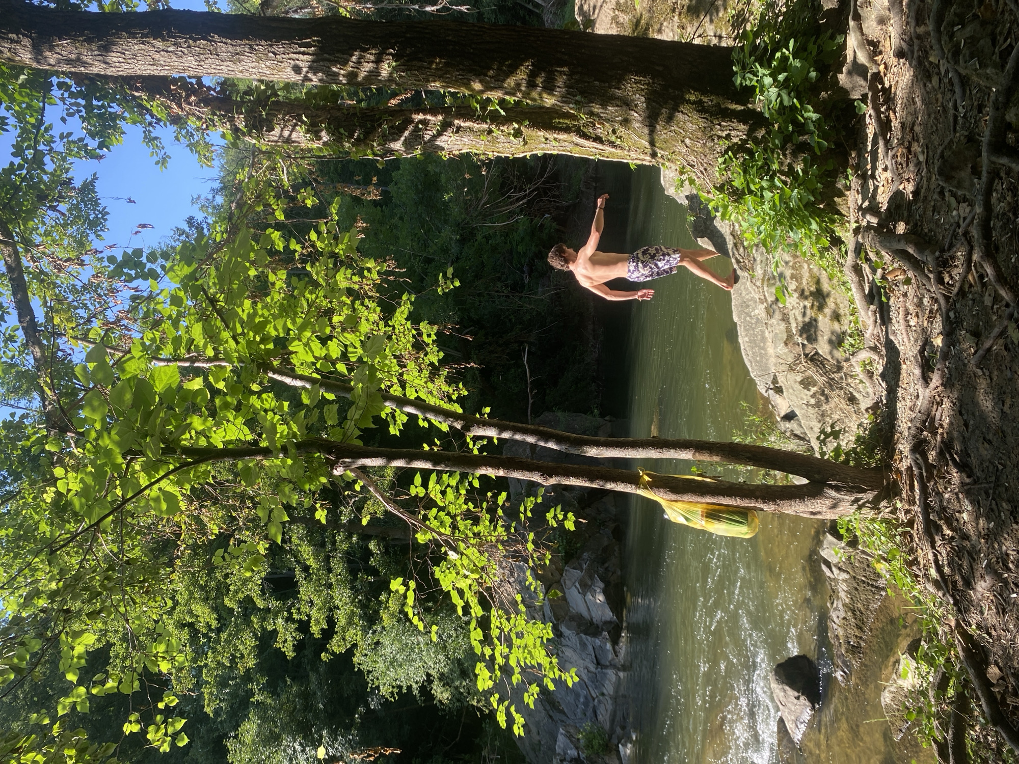 A young man, bare-chested and wearing patterned swim trunks, stands on a rocky bank beside a river in Debnevo, Bulgaria. He is facing the water, with his left arm extended outward and his right hand at his side. The river flows with moderate current, with some rocks visible in its shallows and along the bank. Lush green trees and foliage surround the scene, with sunlight filtering through the leaves, creating dappled shadows on the ground. A bright yellow bag hangs from a tree near the river's edge. The overall impression is of a serene, natural setting.