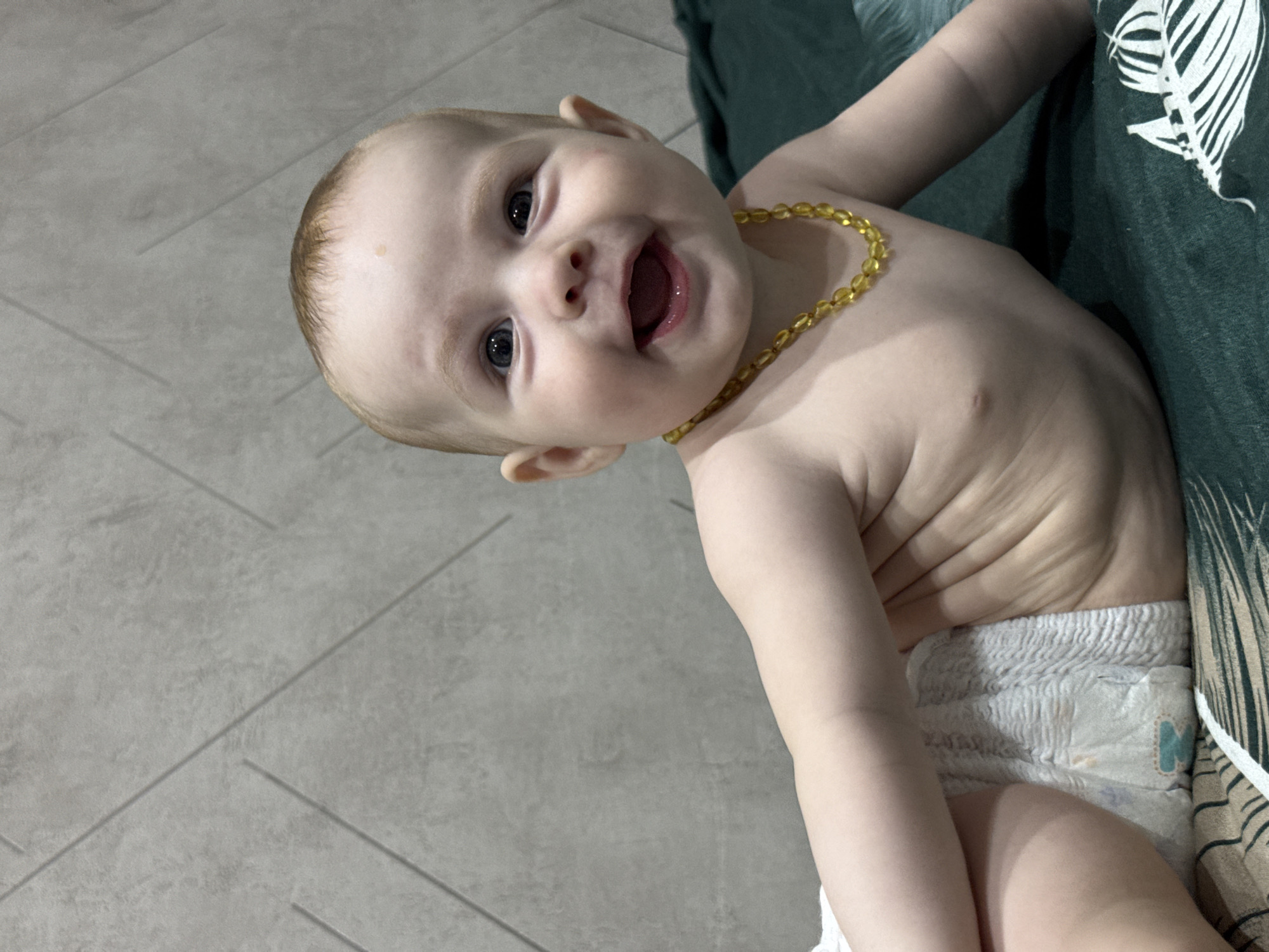 A close-up, chest-up portrait shows a baby girl lying on her stomach on a dark green textured surface, possibly a blanket or bedspread, with a patterned fabric visible at the bottom. The baby is wearing a diaper and a yellow beaded necklace. She is looking towards the camera with a wide, joyful smile, her mouth open and her eyes bright and dark. Her skin is fair, and she has light brown hair. The background is a textured grey wall with a subtle geometric pattern. The lighting is soft, highlighting the baby's features and creating a warm, intimate atmosphere. The scene appears to be indoors. There is no information in the image to determine the location in Noviy, Russia.