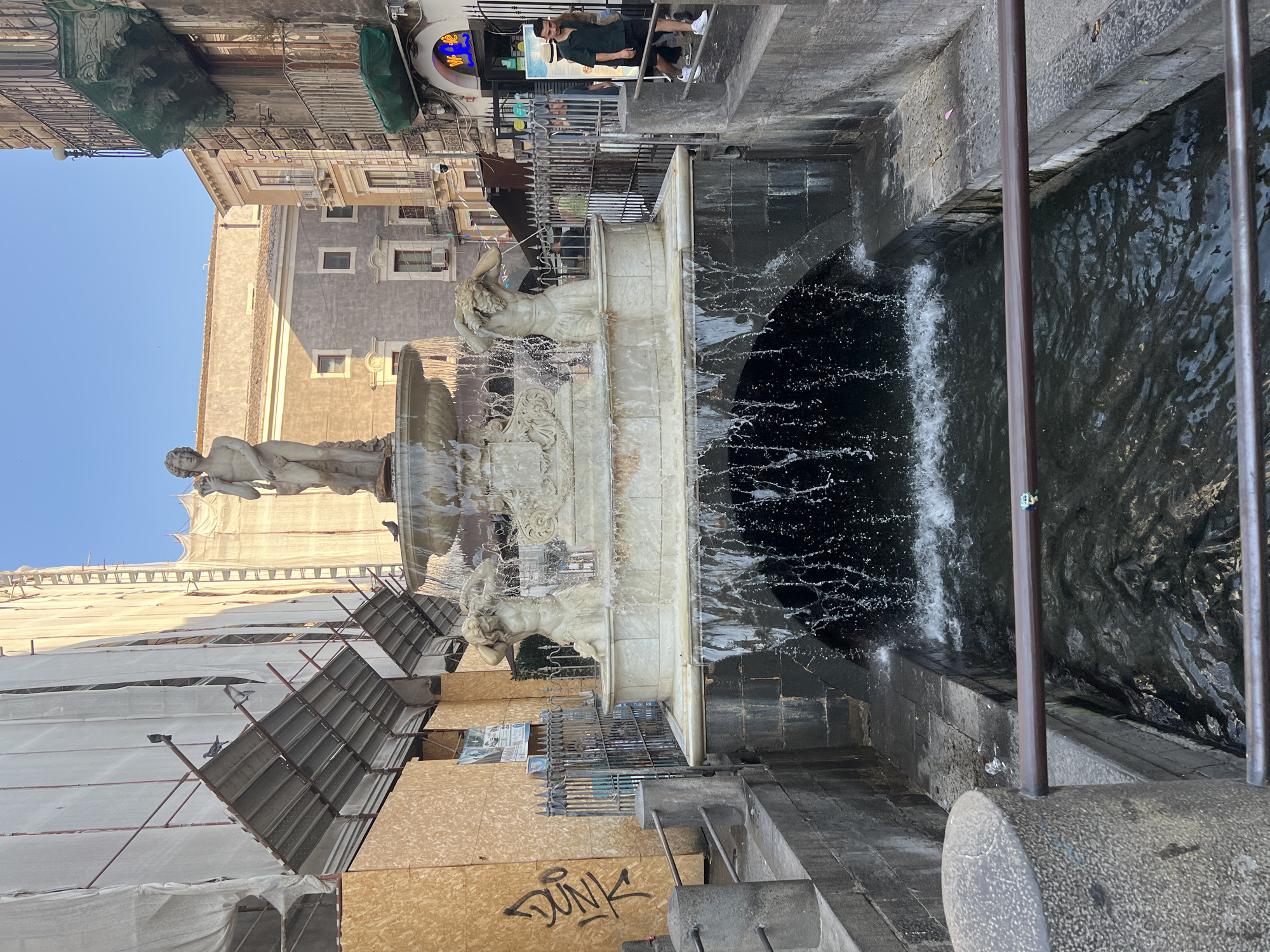 The image shows a public fountain in an urban setting, likely Catania, Italy, given the context. The fountain is ornate, made of white stone, and features sculptures of mythological figures, with water actively flowing and splashing. The water cascades into a dark basin, with visible ripples and spray.
In the background, buildings are visible. One building on the left is covered in scaffolding and white tarpaulin, suggesting ongoing renovation or construction. Another building on the right appears older, with a more traditional architectural style. There are people present, one standing near the top of the fountain and another seated on a ledge. The scene is lit by natural sunlight, indicating it's daytime. The sky is clear and blue.
The perspective of the image is slightly tilted, giving a dynamic feel to the scene. The overall impression is of a lively public space with historical elements. There is some graffiti visible on a wooden panel in the foreground, with the word "DUNK" clearly legible. A neon sign with blue and yellow elements is also visible in the upper right portion of the image.