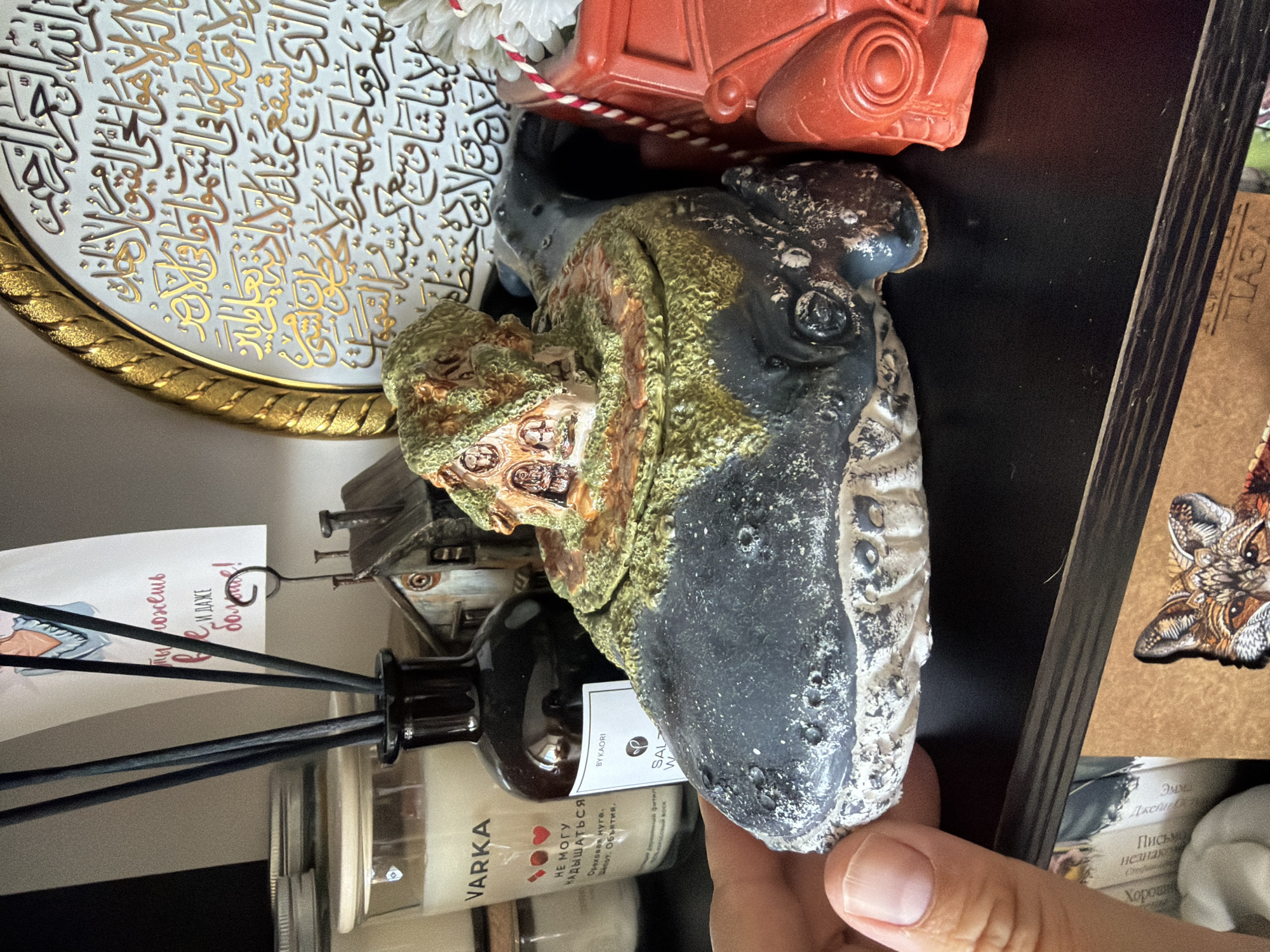 An indoor shot shows a black shelf with various decorative items. In the foreground, a hand is holding a dark grey, whale-shaped ceramic pot with a miniature house on top, covered in moss and vegetation. To the right of the whale pot is a red toy car. Behind these objects, on the shelf, are a glass jar with a white lid, a dark brown bottle with a label that reads "BY KAORI", a small rustic house figurine, and a framed piece of art with Arabic calligraphy. To the left of the whale pot, a card with a drawing and Russian text is held by a clip. The background includes other shelves with more items. The scene is likely a home or a shop display. The user mentions the location is Chaikovsky, Russia, but there are no specific visual cues in the image to confirm this.
