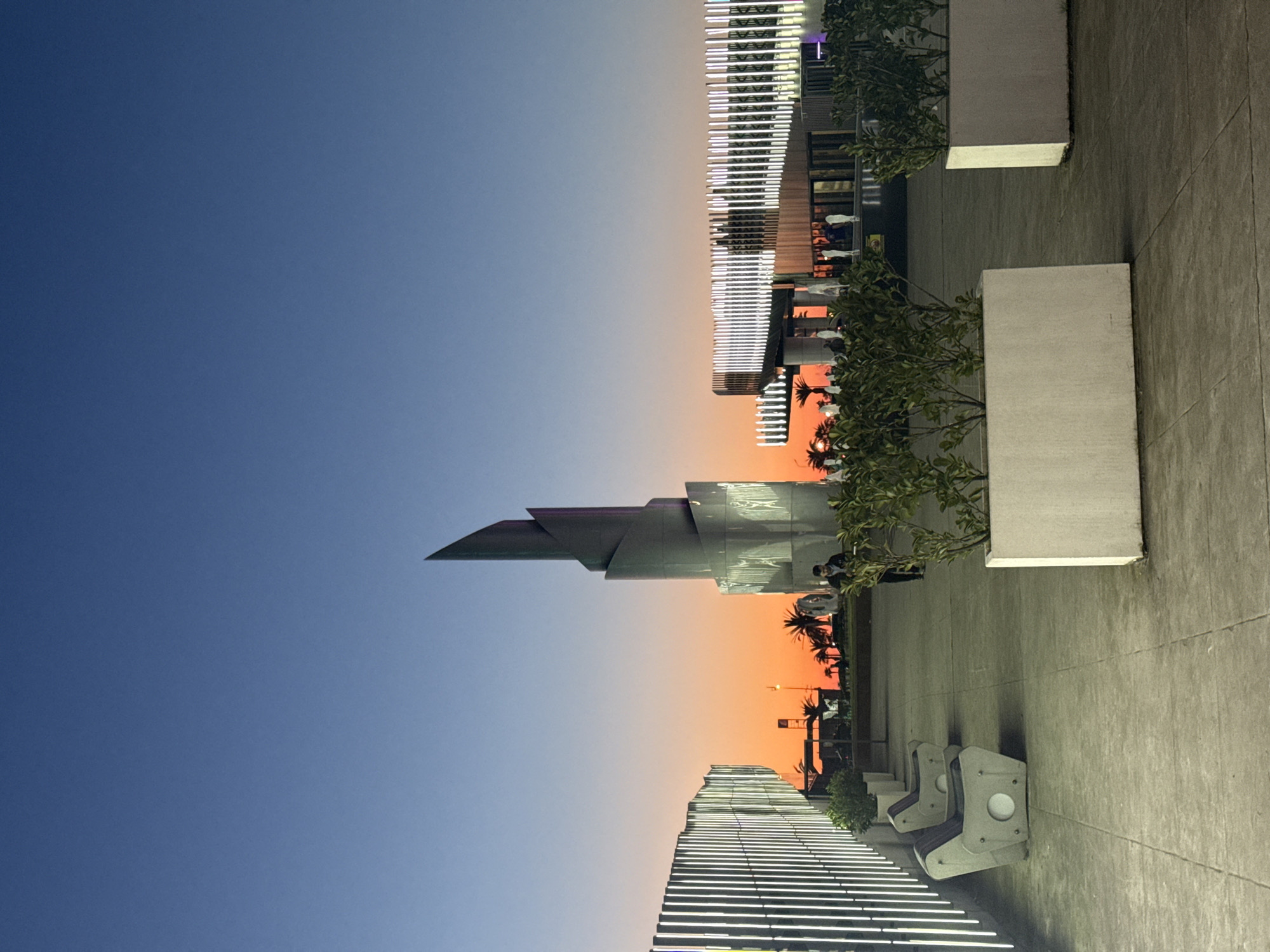 Here is a description of the image:
The image depicts a twilight cityscape in Batumi, Georgia, notable for its modern architecture set against a gradient sky shifting from blue to orange. The most prominent building is the Alphabet Tower, characterized by its unique spiral design. Below the tower is a stepped building featuring horizontal lighting. Along the right side of the image is a tiled walkway with planters and benches. A few people are visible in silhouette. The scene captures a blend of urban design and natural light.