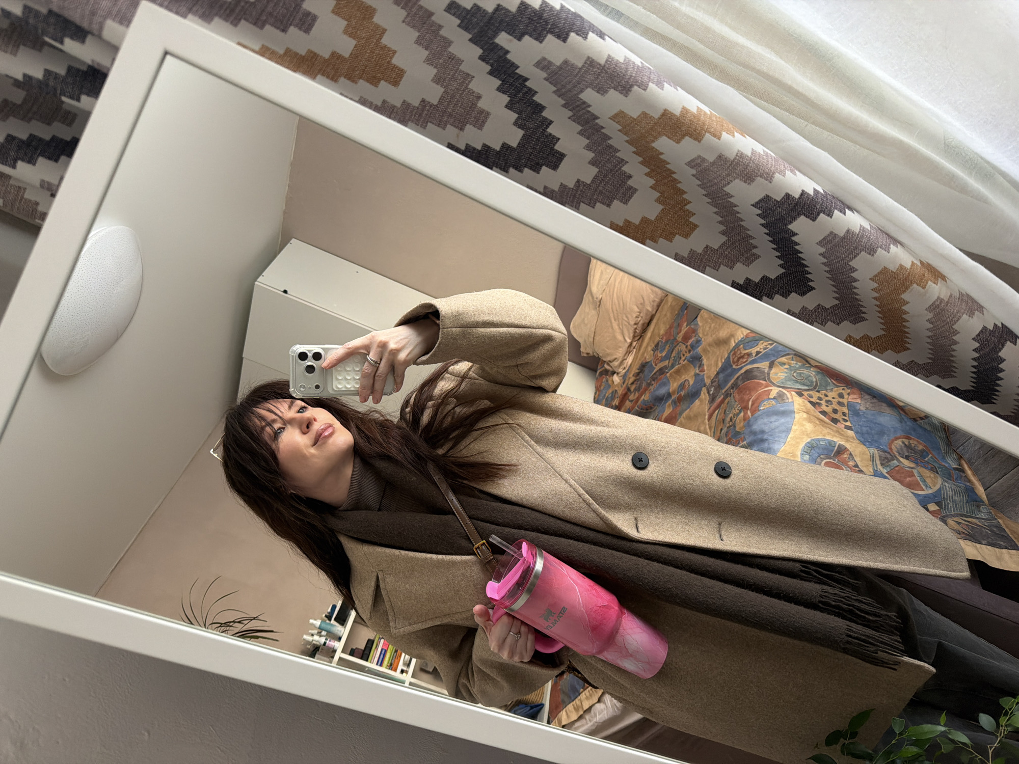 A female individual is depicted in a full-length mirror selfie taken within an indoor residential setting in Perm, Russia. She has long, dark brown hair and is wearing a light brown double-breasted coat, a dark brown turtleneck, and a dark brown fringed scarf. Her left hand holds a large, pink and white marble-patterned Stanley Quencher H2.0 FlowState tumbler equipped with a handle, a dark brown shoulder strap, and a straw. A silver ring is visible on her left ring finger. She holds a light-colored smartphone with a textured white case in her raised right hand, capturing the reflection. The individual is looking slightly to her right with a subtle smile.
The reflected environment includes a light-colored wall on the left, featuring a white shelving unit stocked with various bottles and books. A houseplant is partially visible at the bottom left. On the right, a bed is visible with a patterned duvet in shades of blue, brown, and orange. Light-colored curtains with a zig-zag pattern in brown, gray, and white are present, alongside a sheer white curtain. An oval-shaped white ceiling light fixture is reflected at the top of the mirror.