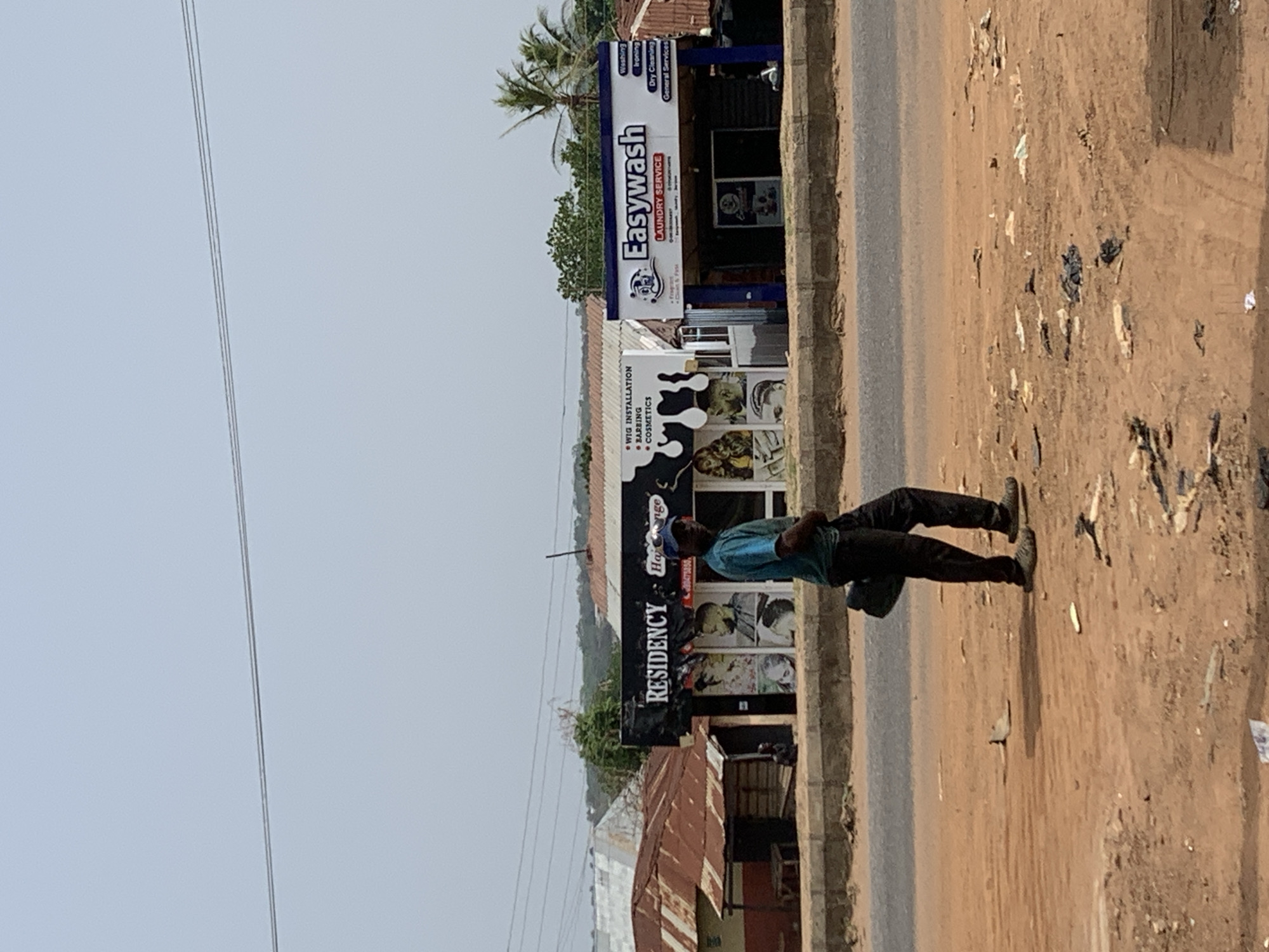 The image depicts an outdoor scene in Ilorin, Nigeria, along a street. A man wearing a blue shirt and dark pants is walking along the right side of the road. To the left of the road are commercial establishments. A business called "RESIDENCY" with signage indicating "WIG INSTALLATION, BARBING, COSMETICS," and another named "Easywash LAUNDRY SERVICE" are visible. A utility pole with wires is seen against a bright sky. Debris is scattered on the unpaved ground beside the road. A palm tree is present near the businesses. Adjacent to the commercial buildings is what looks like the edge of residential housing with tin roofs.