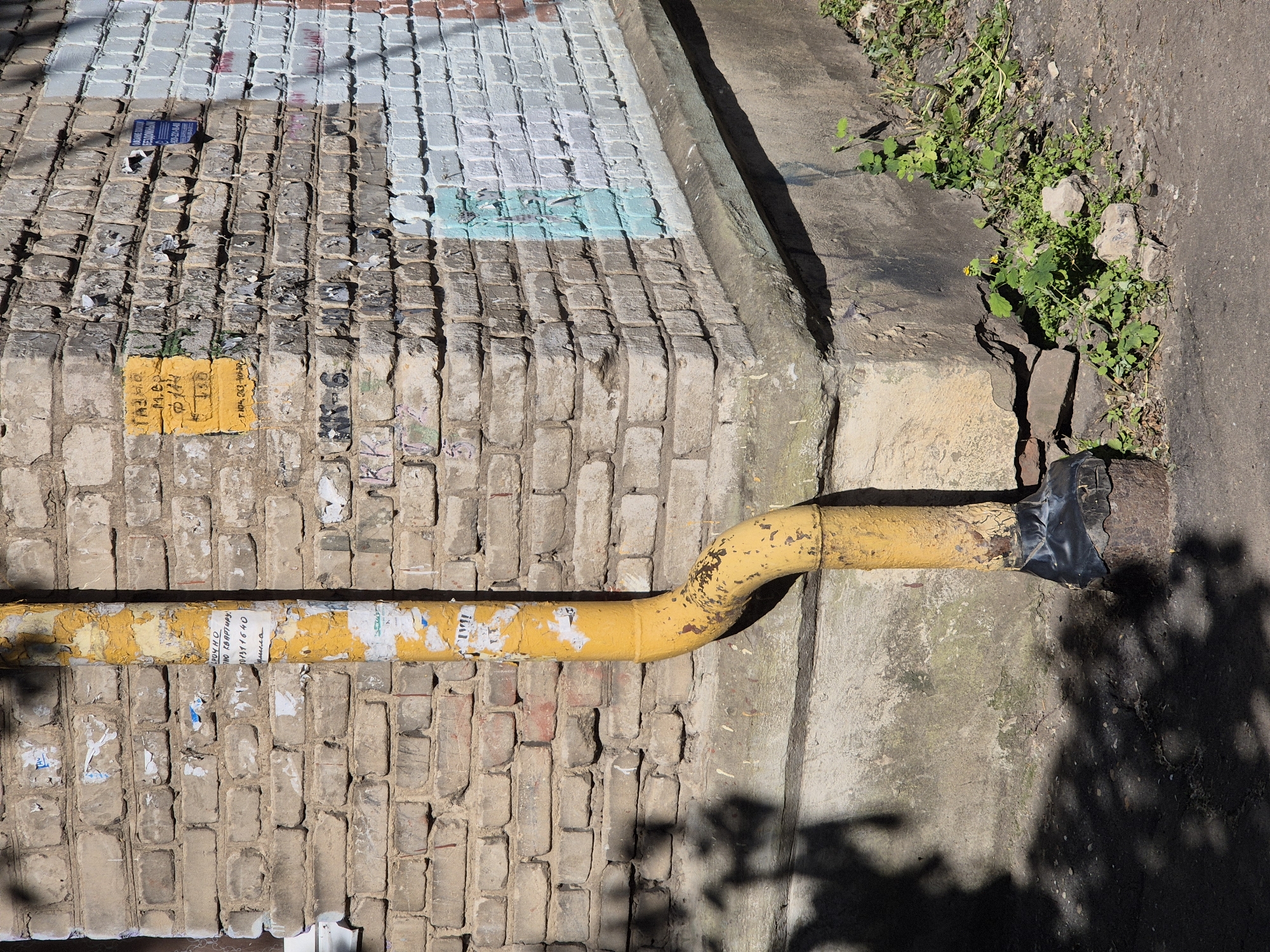 A downspout, painted yellow and peeling, runs vertically down the corner of a brick building. The building's facade is constructed of weathered, light-brown bricks, with some sections painted white, light blue, and red. Various pieces of torn paper and graffiti are visible on the walls. At the base of the downspout, a rusty metal pipe is capped with black tape. The ground in front of the building is a mix of asphalt and dirt, with patches of green weeds and small yellow wildflowers growing near the foundation. Shadows from unseen foliage are cast on the wall and ground.