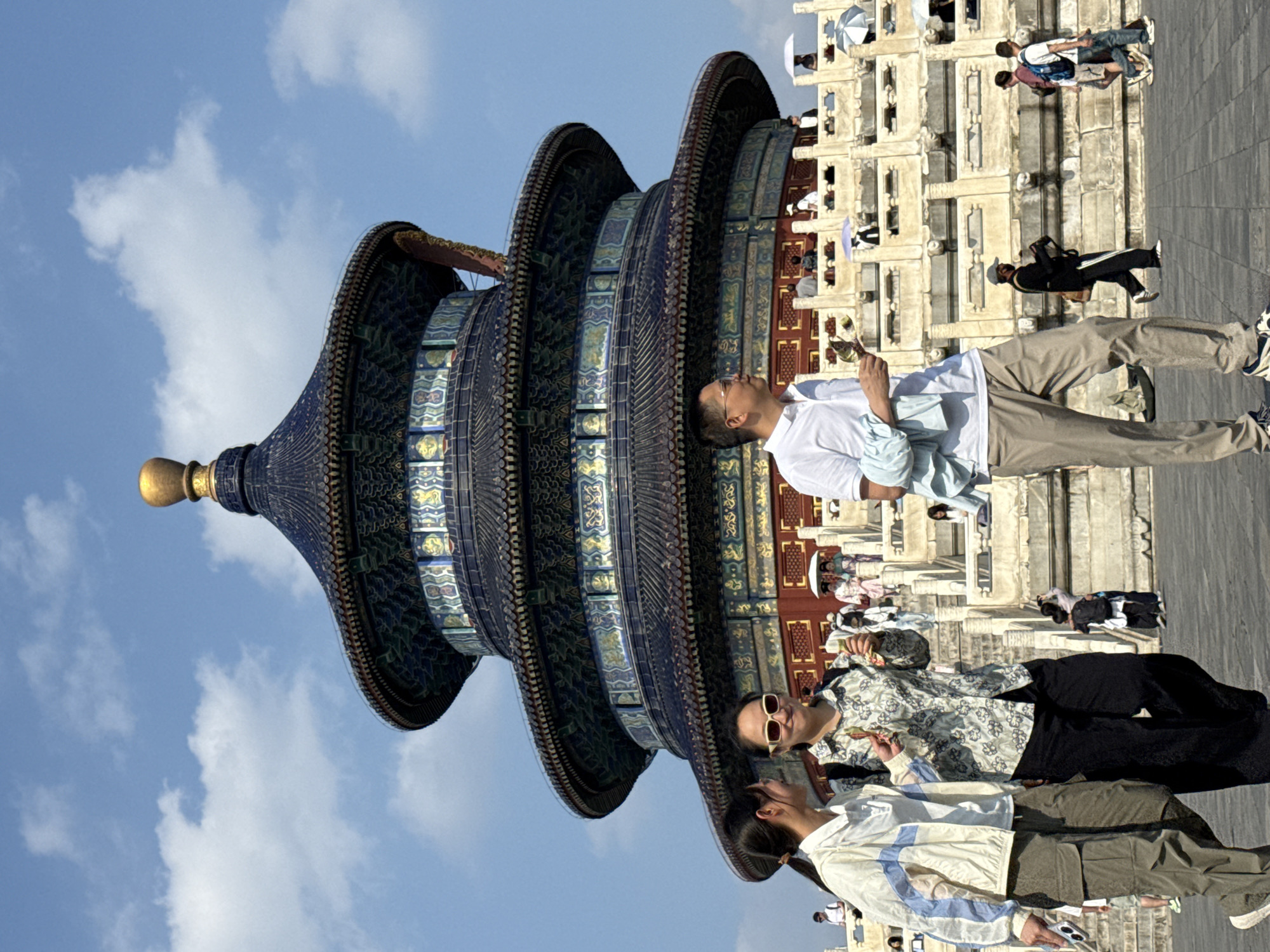 The image depicts the Hall of Prayer for Good Harvests, a key structure within the Temple of Heaven complex in Dongcheng District, China. The iconic multi-tiered, circular building with a dark blue, tiled roof and a golden finial is centrally located. In the foreground, three individuals are walking. The two women on the left are in focus, with one wearing a light-colored jacket and cargo pants, and the other in a floral-patterned shirt and dark pants. Both appear to be holding snacks. A man in a white polo shirt and beige pants is walking in front of them, to the right. Behind these individuals, a wide stone staircase leads up to a terrace where more people are visible, some holding umbrellas. The sky is blue with scattered clouds, suggesting a sunny day. The overall scene captures a moment of tourism and exploration at this significant historical site.