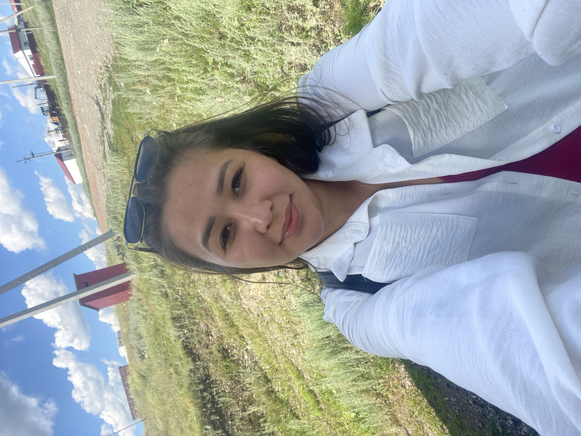 A woman takes a selfie outdoors with a red outhouse and buildings in the background. She is wearing a white button-down shirt and sunglasses are on top of her head. The landscape is grassy with a dirt road. The sky is blue with white clouds.