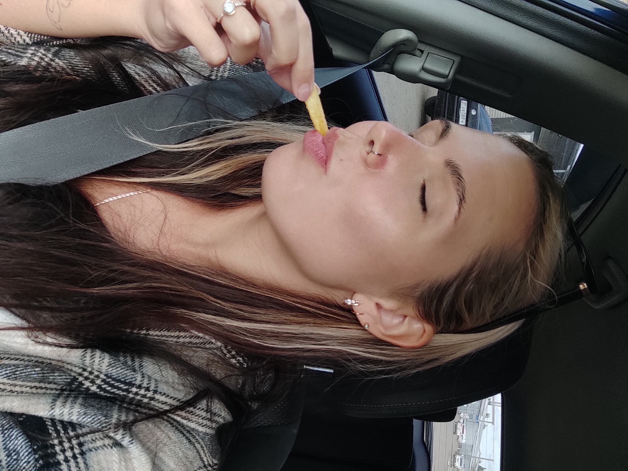A close-up, portrait-oriented photograph captures a young woman inside a car, seemingly in Chaikovsky, Russia. The woman, with her head tilted back and eyes closed, is in the process of eating a french fry, held to her lips by an unseen hand. She wears a plaid pattern garment in grey, white, and black, with a seatbelt diagonally crossing her chest. Her long, dark hair with prominent blonde highlights frames her face. Notable features include a silver hoop nose ring on her left nostril, a thin chain necklace, and multiple earrings on her right ear, including a prominent three-stone stud and smaller hoops. Sunglasses are hooked over her right ear. The hand holding the fry has a ring with a large, light-colored stone on the ring finger.
The car's interior details, such as a black headrest and parts of the overhead console, are visible. Through the car's rearview mirror, an outdoor scene with buildings and what appears to be a street or parking lot can be seen. Faint text, "21 BO89A," resembling a license plate or vehicle tag, is visible in the reflection within the mirror. The bright lighting suggests it is daytime. The woman's closed eyes and open mouth imply a moment of relaxed enjoyment while eating.
