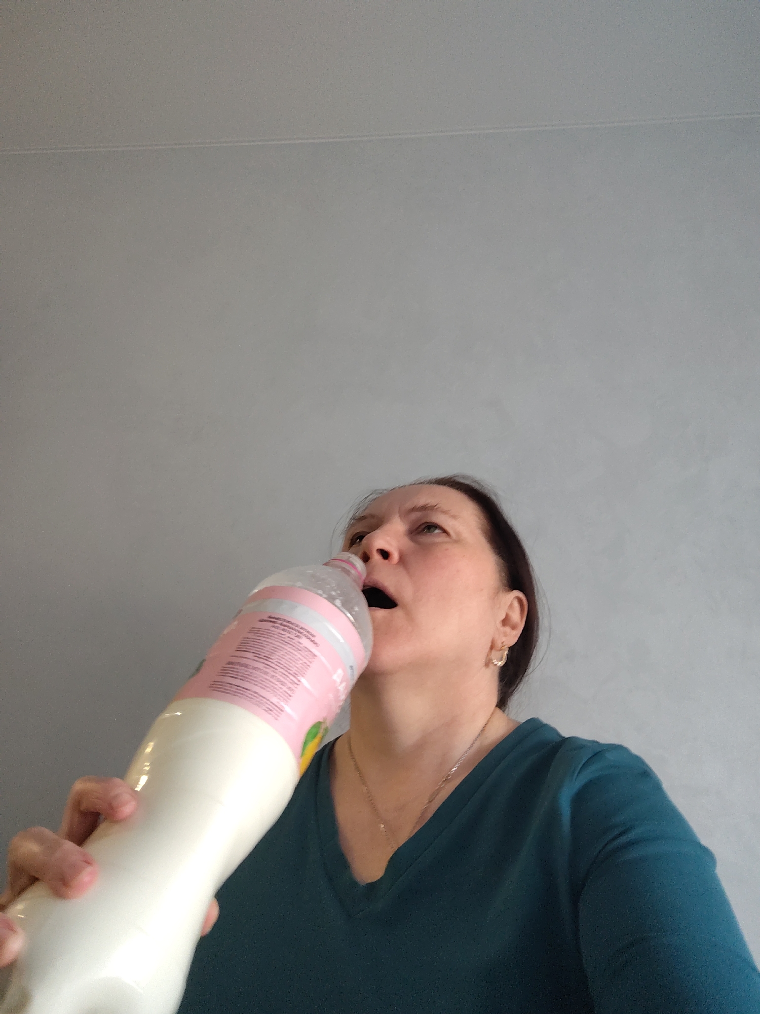 A middle-aged Caucasian woman is drinking from a large, clear plastic bottle. The bottle contains a white liquid, possibly milk, and has a pink label with text in Russian. The woman has brown hair and is wearing a teal V-neck shirt and a silver necklace. She has a gold hoop earring in her left ear. The background is a plain gray wall. There is no evidence to suggest that this image was taken in the city of Neftekamsk, Russia.