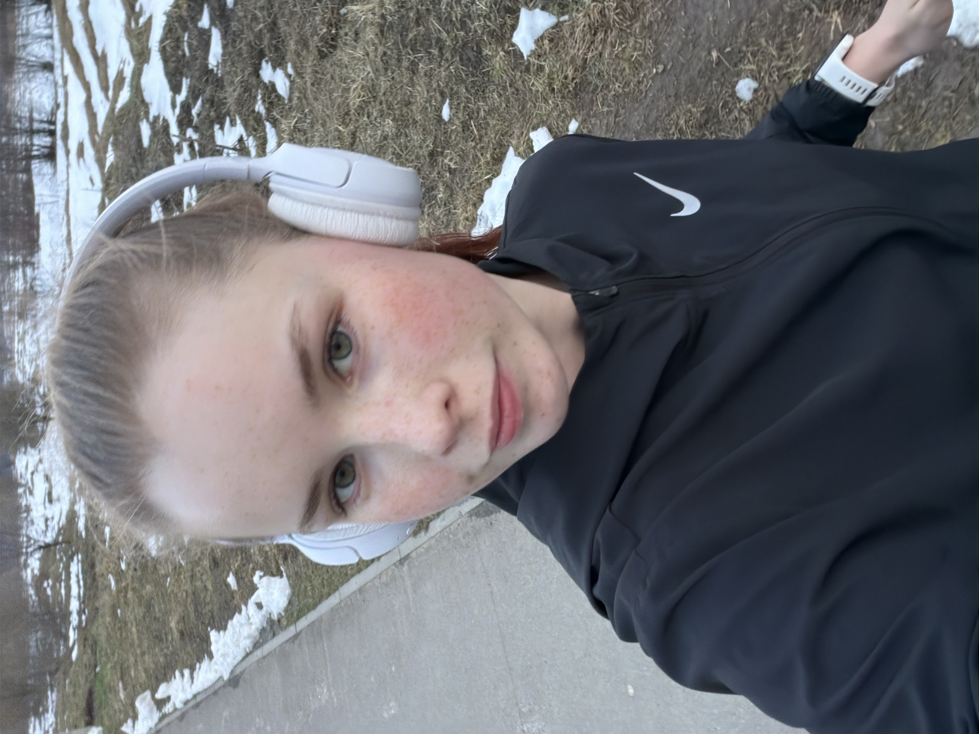 This is a vertical selfie of a young woman outdoors in Berezniki, Russia, likely during late winter or early spring. The subject is a Caucasian woman with light skin, numerous reddish freckles across her cheeks and nose, and light green eyes. Her light brown hair is pulled back, and she is looking directly at the camera with a neutral to slightly smiling expression.
She is wearing white over-ear headphones and a black athletic jacket with a prominent white Nike swoosh logo on the left chest. A white smartwatch or fitness tracker is visible on her left wrist.
The setting is an outdoor area featuring patches of melting snow scattered on dry, brown grass or ground, indicating a cool, transitional season. Bare trees are visible in the background, further supporting the late winter or early spring timeframe. A grey concrete path or sidewalk runs along the lower and right portions of the image, suggesting she is on a pathway. The lighting suggests it is daytime. The overall scene depicts an individual engaged in an outdoor activity, possibly a walk or run.