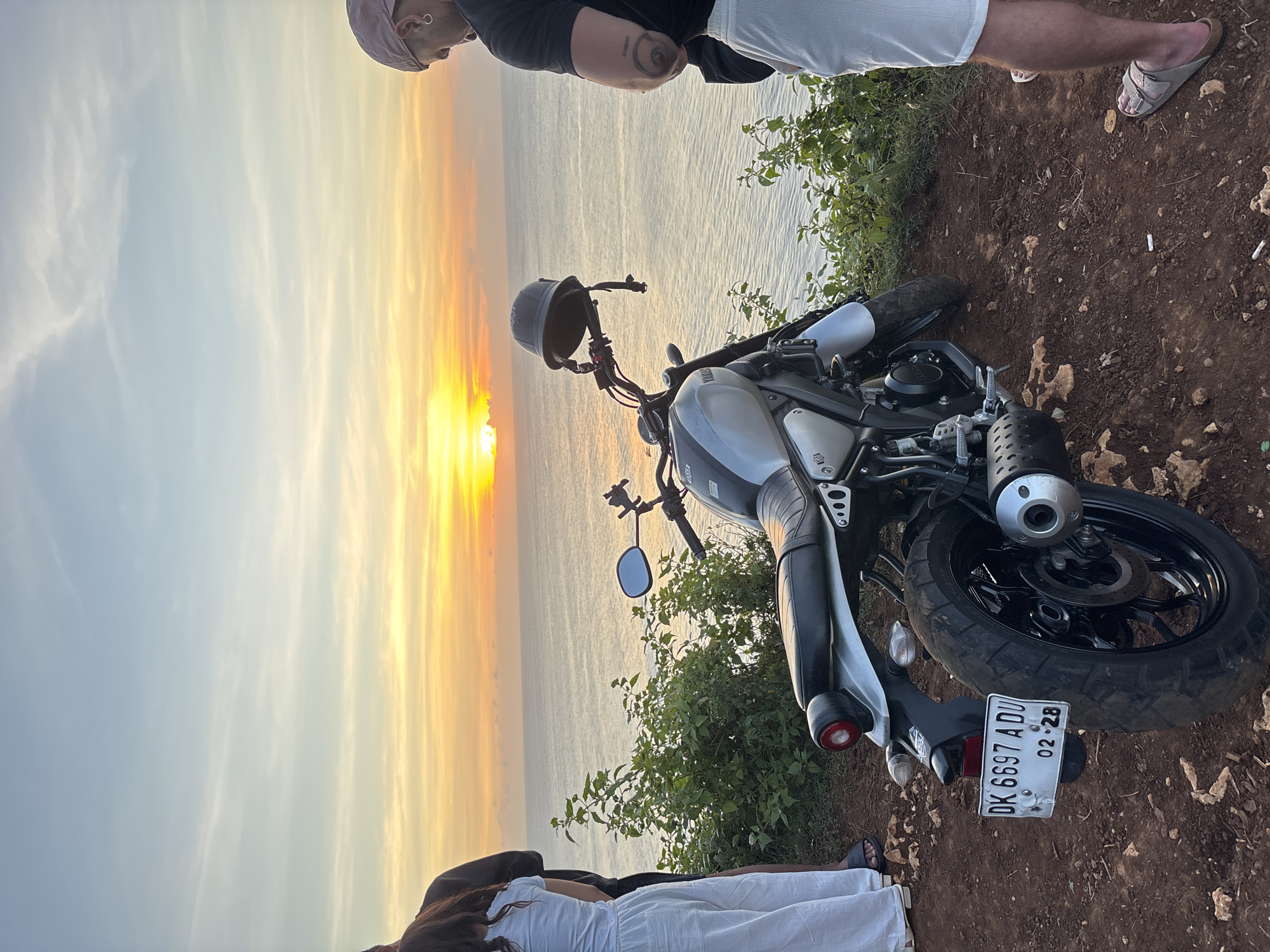Captured from a high vantage point in Kuta Selatan, Indonesia, the image displays a motorcycle parked on a dirt verge overlooking the ocean, set against a sunset sky. The motorcycle is a silver Yamaha with a black seat and handlebars. A black helmet is placed on the handlebars. The license plate reads "DK 6697 ADU 02.28". To the left of the motorcycle, a female figure in a white dress and dark hair is partially visible. To the right, two other figures are partly captured: one wearing a black t-shirt and white shorts, and the other in white shorts and wearing sandals. The dirt verge has greenery including grass and small plants. The ocean extends to the horizon, where the sun is setting, casting a bright orange and yellow glow. The sky is a mix of blue, orange, and yellow hues.