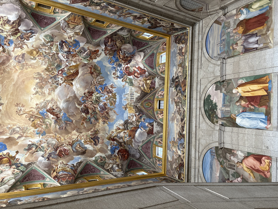 The image depicts a grand, ornate interior space within the Royal Monastery of San Lorenzo de El Escorial, San Lorenzo de El Escorial, Spain. The scene is dominated by extensive fresco painting covering the vaulted ceiling and upper walls, with additional murals on the lower wall sections.
The vaulted ceiling is covered by a vast Baroque fresco, portraying a dynamic celestial scene. Hundreds of human and angelic figures are depicted in various active poses, ascending through swirling clouds towards a central, luminous light source. Elements such as a cross are visible being carried aloft. The color palette is vibrant, featuring prominent blues, whites, reds, greens, and gold accents.
Below the main ceiling fresco, the upper wall sections feature smaller, horizontally oriented rectangular murals. These murals depict historical or battle scenes, including numerous human figures, cavalry, and detailed landscapes, rendered in a more subdued, earthy color scheme. These painted sections are framed by ornate architectural elements and gold trim. Several small, rectangular windows are visible within these upper painted areas, allowing natural light.
The lower section of the wall is constructed from large, visible cut stone blocks. Embedded within this stonework are three distinct, large arched murals. The leftmost mural, partially visible, shows a cloaked figure kneeling before another against a landscape background. The central mural depicts a standing figure in a blue robe conversing with two cloaked figures, one in red and one in gold, beneath a tree with distant architectural elements. The rightmost mural presents a group of at least four standing figures in various cloaks and robes, engaged in interaction within a landscape that includes a distant city.
The overall perspective is upwards, emphasizing the immense scale and intricate detail of the integrated ceiling and wall artistry.