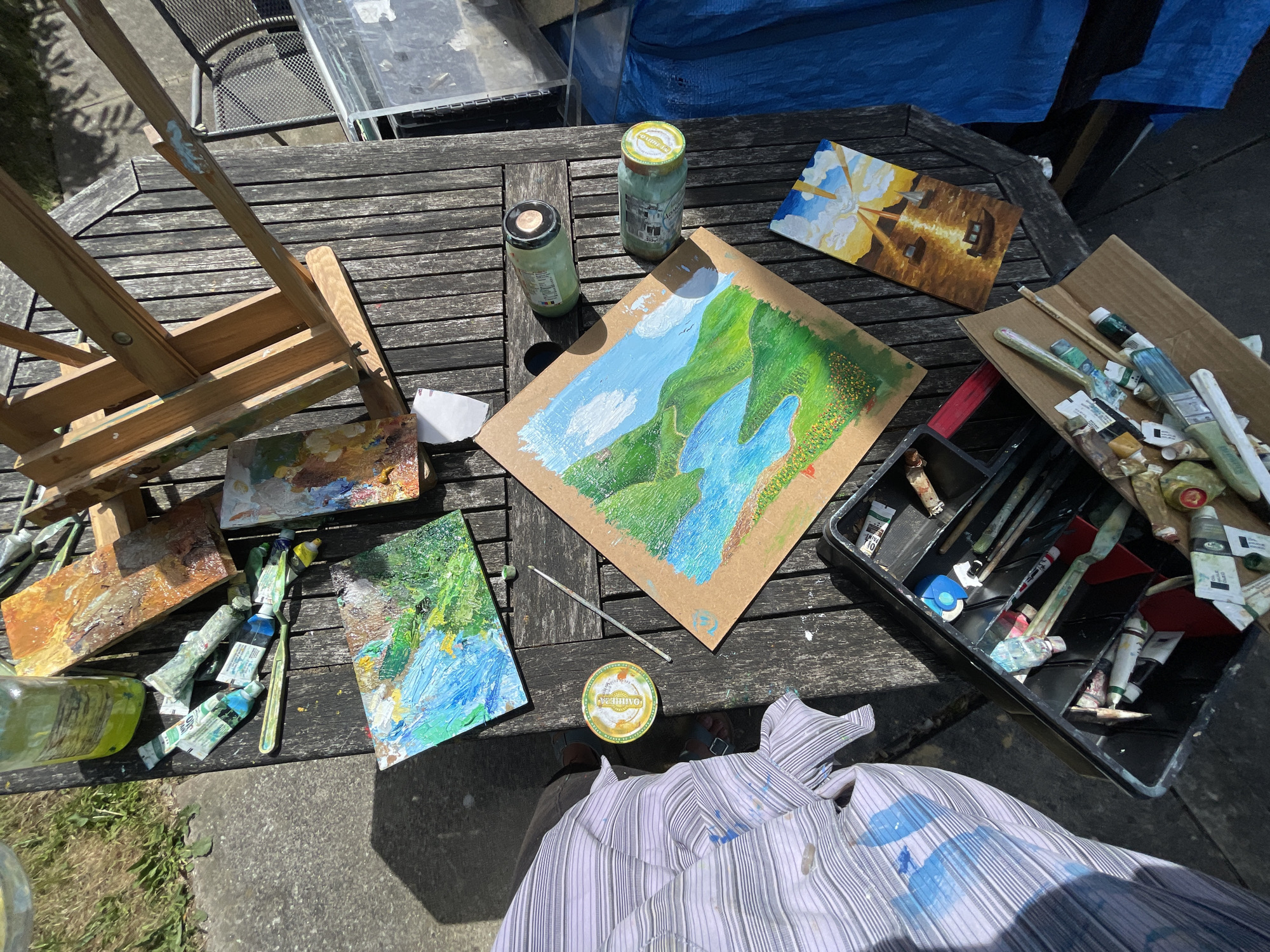 The image depicts an outdoor painting setup on a wooden table in Maidstone, United Kingdom. An easel sits at the table's edge, alongside a palette with dried paint. Several partially completed paintings are scattered on the table, depicting landscapes and buildings. Two jars with lids and tubes of paint are also present. A black container holds an assortment of paintbrushes and painting tools. A person wearing a paint-splattered shirt is positioned in front of the table. The setting includes a partial view of pavement and lawn in Maidstone, United Kingdom.