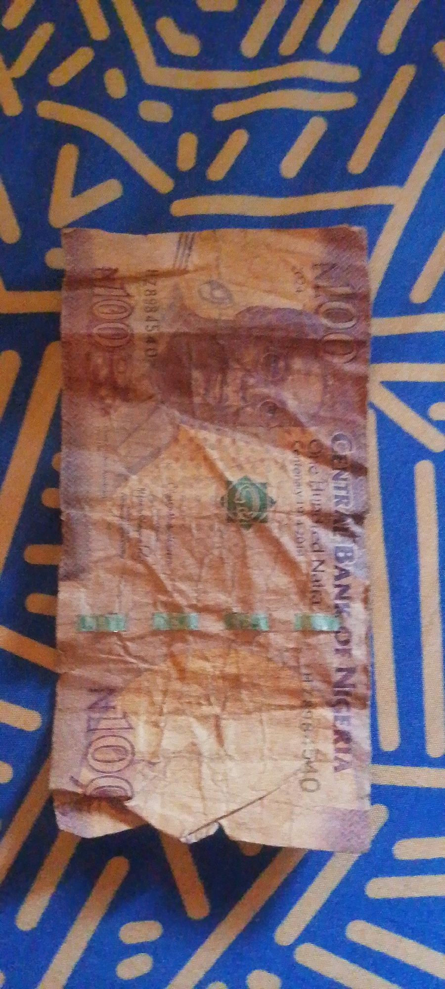 The image shows a crumpled Nigerian 100 Naira banknote placed on a blue and yellow patterned fabric. The banknote is folded in half vertically and appears to be made of paper. The visible text on the banknote includes "100", "CENTRAL BANK OF NIGERIA", "One hundred Naira", and a serial number "#289845407". There are also faint images and symbols visible on the banknote, including what appears to be a green emblem. The fabric it is lying on has a repeating pattern of yellow lines and dots on a blue background, resembling a traditional textile or a tablecloth. The lighting in the image suggests it was taken indoors, possibly in daylight. The overall scene is simple, focusing on the banknote as the primary object. There are no people or clear indications of a specific event taking place. The location context provided is Jalingo, Nigeria, which is consistent with the currency shown.