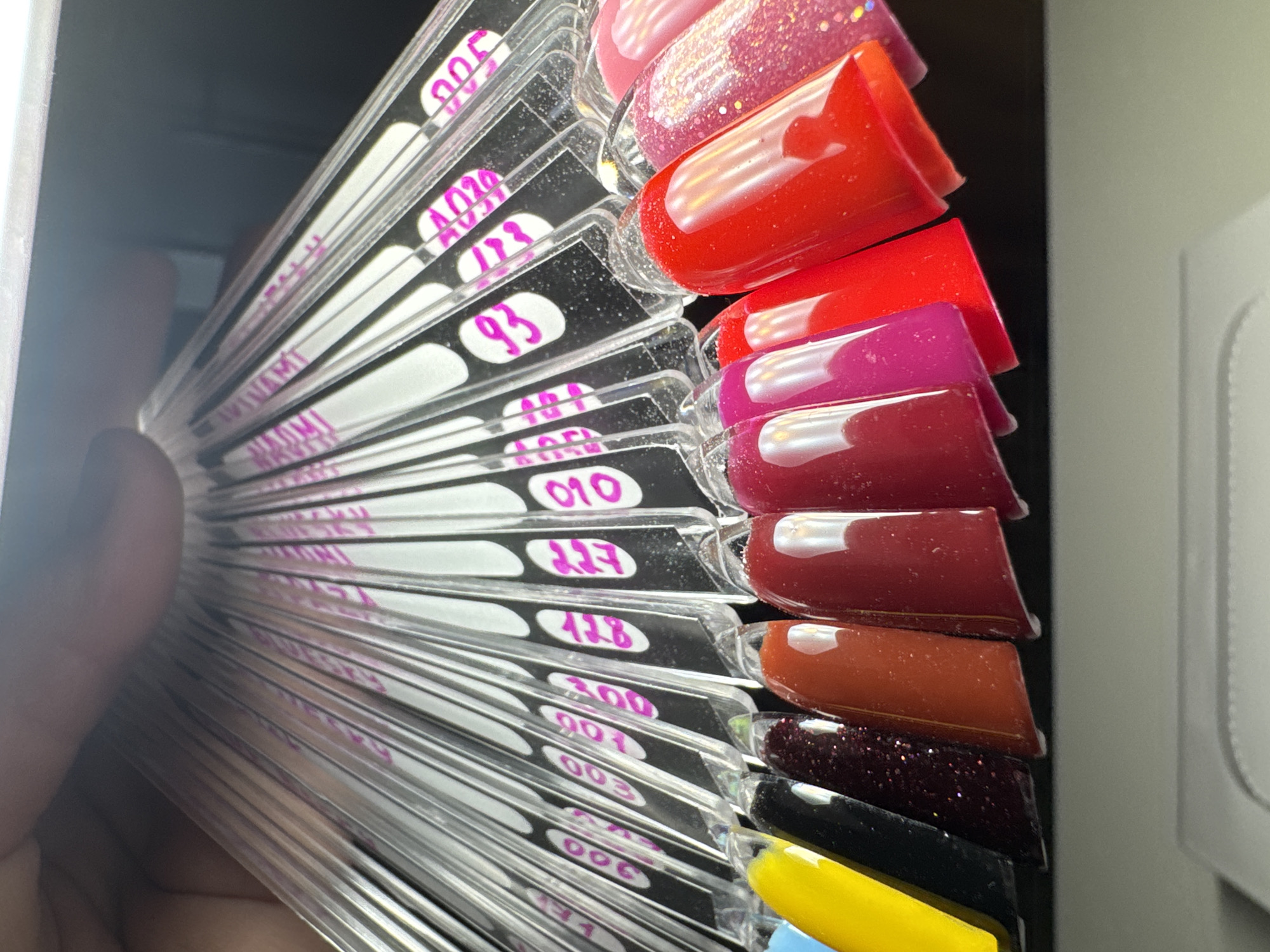 This media file captures a close-up, angled view of a fan-like display of nail polish color swatches being held by a hand. The swatches are made of clear plastic, with each tip painted a different color and finish, arranged in a fanned-out pattern.
**Subjects:** The primary subjects are numerous nail polish color swatches and a person's hand holding them from the left side of the frame. The swatches showcase a variety of colors, including vibrant reds, fuchsia, burgundy, orange-red, black, yellow, and what appears to be a light blue at the very bottom. Some polishes have a glossy, solid finish, while others feature glitter or sparkle, such as a pink glitter and a dark red/burgundy glitter.
**Activity or event taking place:** The image depicts the act of presenting or selecting nail polish colors from a sample collection. The fanned arrangement allows for easy viewing of multiple options.
**Notable details:** The nail polish samples are attached to clear plastic sticks, which have white oval labels against a black background on their handles. Hand-written numbers and some text are visible on these labels, mostly in purple marker. Visible numbers include 005, A020, 123, 93, 010, 127, 128, 300, 001, 003, 006, and 171. Partially visible text fragments like "MAMY" and "SROMT" or similar illegible words are also present on some labels. The background is dimly lit and blurry on the left, possibly an indoor setting. To the right, a light gray or white, possibly textured, object is visible, hinting at a wall or piece of furniture. No specific time of day or weather is discernible from the image. The general location context provided is Chaikovsky, Russia.