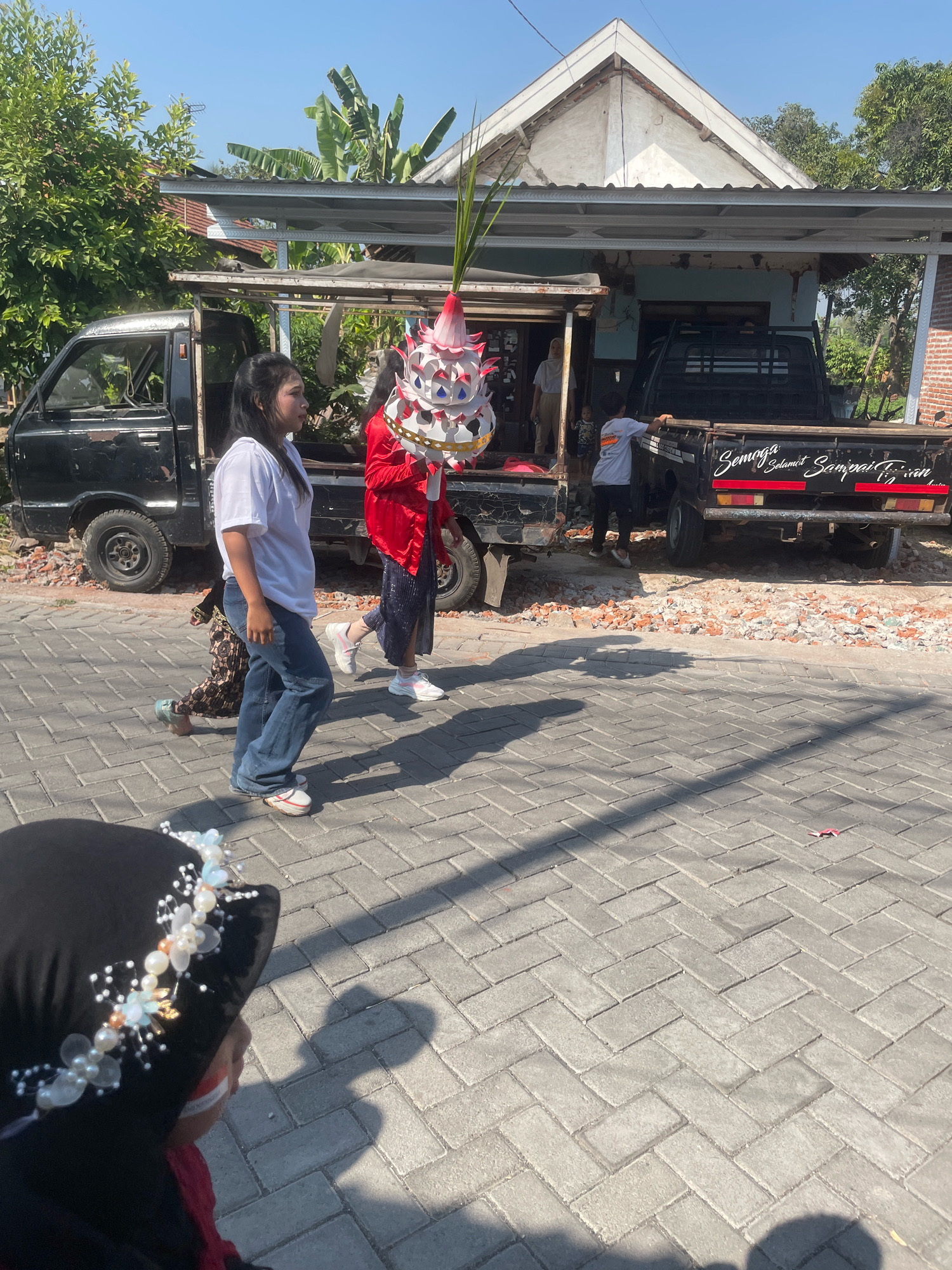In the city of Kebon Waris, Indonesia, on a sunny day, a group of people are participating in a procession. Two black pickup trucks, one with its rear facing the camera and the other on the left side of the image, are parked near a building. The building has a white, triangular roof and appears to be under construction or renovation. A woman in a white t-shirt and blue jeans walks from left to right, carrying a child who is partially obscured. Another woman in a red jacket and sparkly skirt is in the center of the frame, holding a decorative, tiered object topped with green leaves. A third person, wearing a grey t-shirt and dark pants, stands near the rear of the pickup truck on the right. The ground is paved with grey interlocking bricks, and rubble is scattered around the base of the trucks. The foreground is dominated by the back of a person wearing a black hijab decorated with pearls and beads, and a glimpse of a child with face paint is visible.