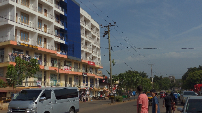 A street scene with a multi-story building on the left, featuring shops on the lower levels and residences above. The building has a modern design with white walls, orange accents, and a blue facade. A utility pole with wires stands near the building. Pedestrians and vehicles, including a silver van, populate the street. Trees line the road, and the sky is partly cloudy.