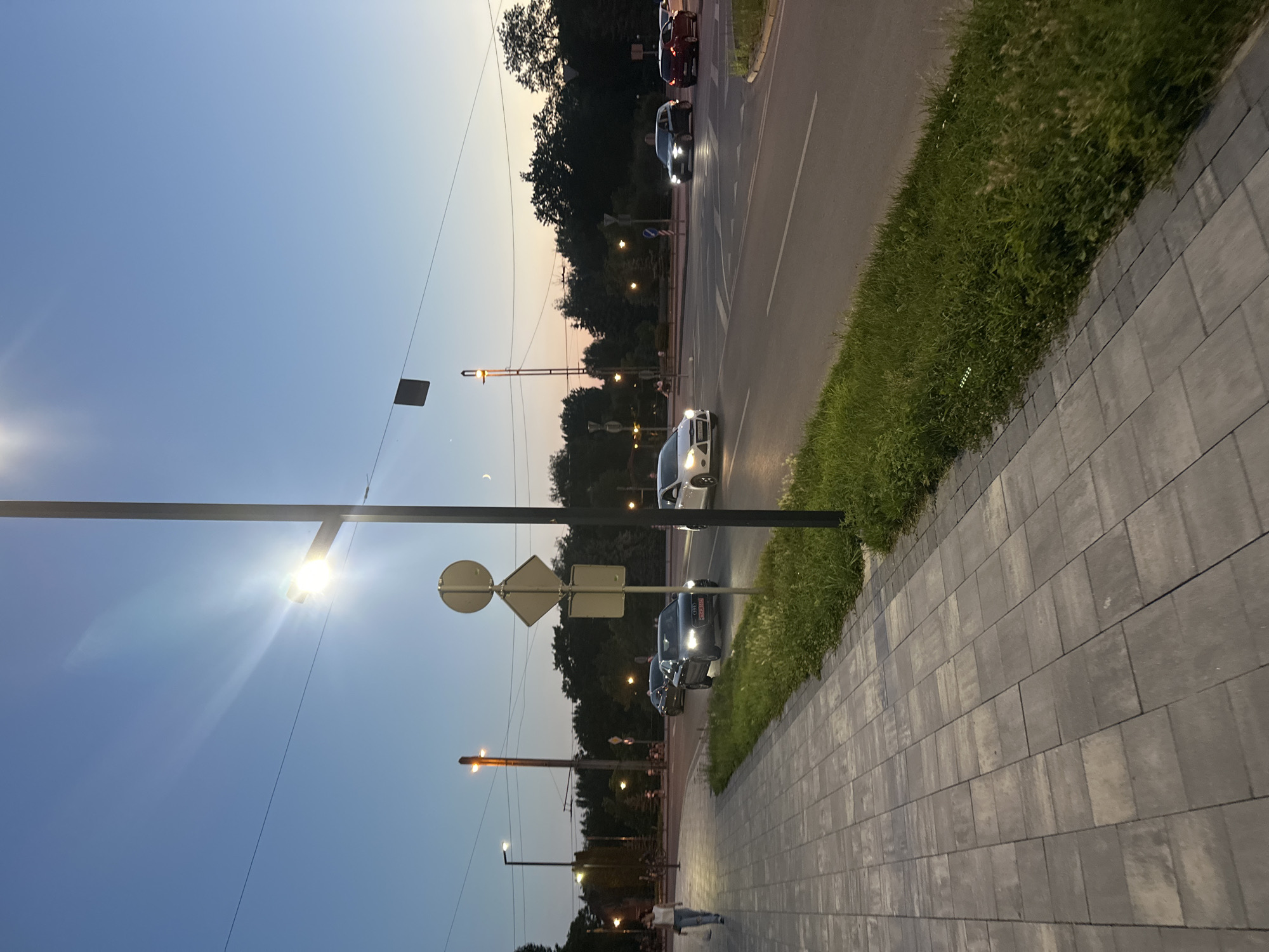 The image captures a twilight street scene in Ternopil, Ukraine. A wide paved sidewalk runs along the left side of the frame, bordered by a strip of lush green grass and then a multi-lane asphalt road. Several cars are visible on the road, some driving and others stopped at an intersection. Streetlights are illuminated, casting a warm glow on the surroundings. A woman is walking on the sidewalk, carrying a shopping bag. In the background, trees line the horizon, and a sliver of a crescent moon is visible in the pale blue sky. Traffic signs are posted at the intersection, and a tall street lamp stands prominently in the foreground. The overall atmosphere is one of a calm evening in a city.
