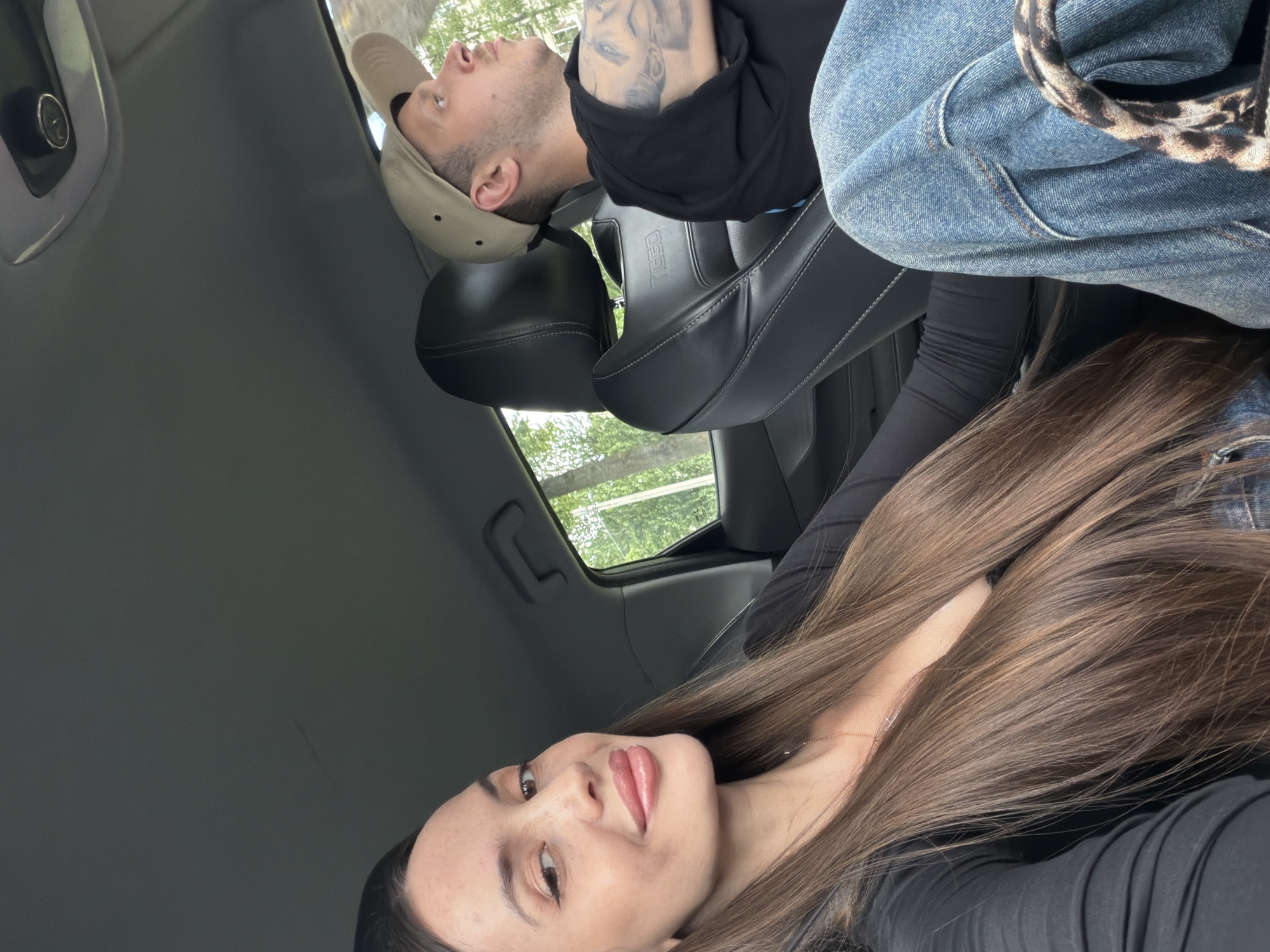 A medium shot shows a woman in the foreground and a man in the background, both inside a car. The woman, positioned on the left, has long brown hair that cascades down her front. She is wearing a black top and a necklace. Her face is turned towards the camera, and she is smiling. Her lips appear to be cosmetically enhanced. The man, on the right, is wearing a black t-shirt, blue jeans, and a beige baseball cap. He is looking out of the car window to the right. His left arm is visible, and it appears to have tattoos. The interior of the car shows black leather seats and the car's headliner. The background outside the car window reveals greenery, suggesting an outdoor location. There is no specific information in the image to indicate the location is in Saint Petersburg, Russia.