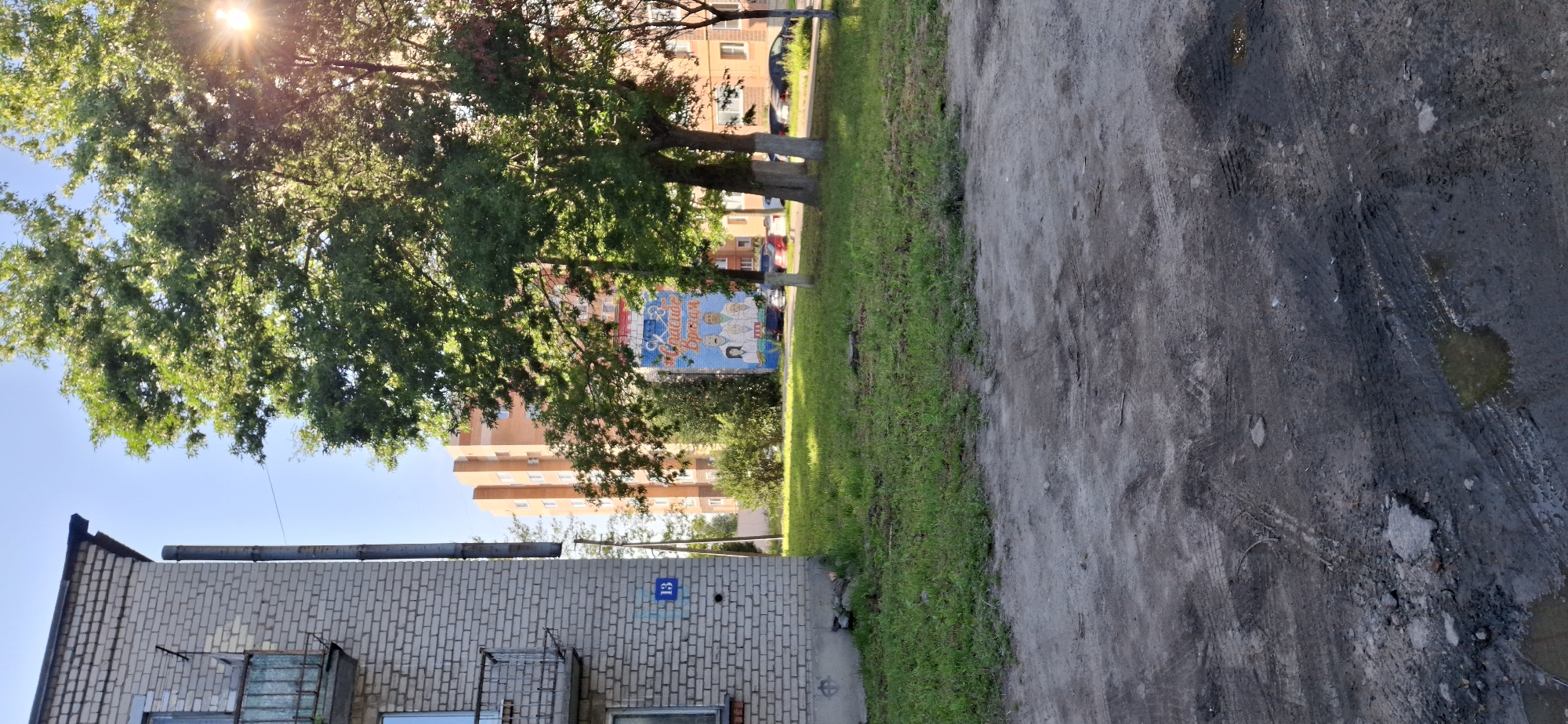 The image shows an exterior view of a residential area on a sunny day. On the left is a section of a light-colored brick apartment building with a balcony and a vertical pipe. A blue sign with the number "13" is affixed to the building. In the background, a taller, orange brick apartment building is visible. In the middle ground, there is a mural on the side of a building depicting several figures, including what appear to be medical professionals, with Russian text above them that reads "Спасибо Врачам" (Thank you Doctors). Green grass covers the ground in front of the buildings, and several trees are present, one with sunlight filtering through its leaves. Cars are parked in the background. The foreground is a rough, dirt and gravel area with tire tracks and some puddles of water, suggesting recent rain or a poorly maintained surface. The scene appears to be located in a residential neighborhood in Telmana, Russia.