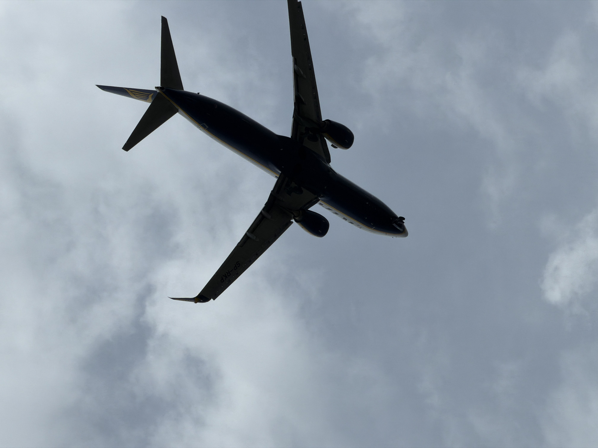An airplane, identified by its livery as belonging to Ryanair, is captured in silhouette against a cloudy sky. The aircraft is seen from below and to the side, with its tail fin, wings, and engines clearly visible. The registration number "SP-RKP" is partially legible on the underside of one wing. The plane appears to be in flight, likely approaching or departing from an airport. The cloudy sky suggests overcast weather conditions.
This image does not contain any information that would allow for its specific location within the city of Burgas, Bulgaria, to be determined.