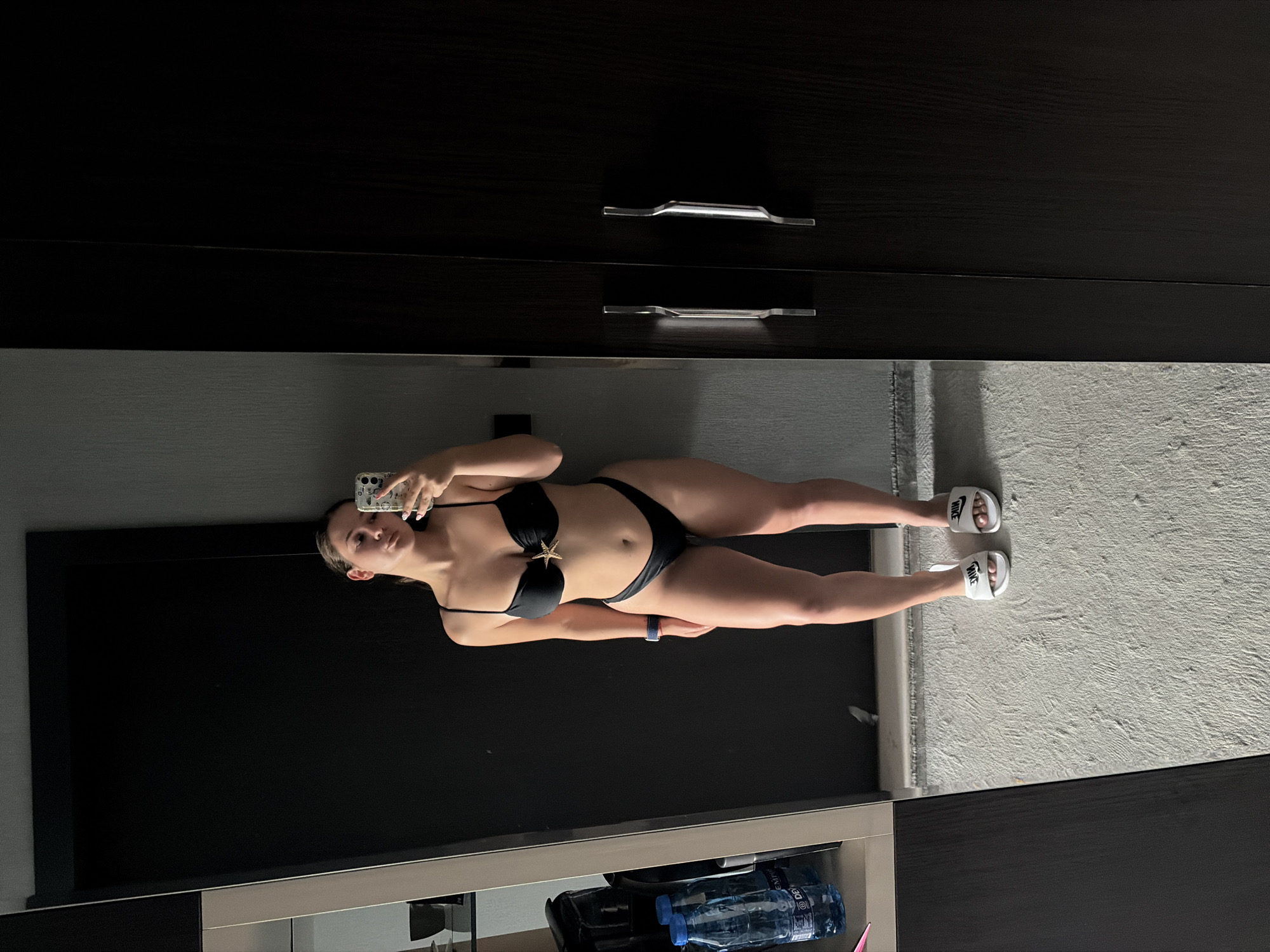 A young woman is taking a full-length mirror selfie in what appears to be a hotel room in Hisarya, Bulgaria. She is wearing a black bikini with a star-shaped embellishment on the top and white Nike slides. She is standing in front of a dark closet and a dark door, with a mirror to her left that reflects part of the room, including a shelf with water bottles and bags. The floor is a light-colored, textured carpet. The overall lighting is dim.
