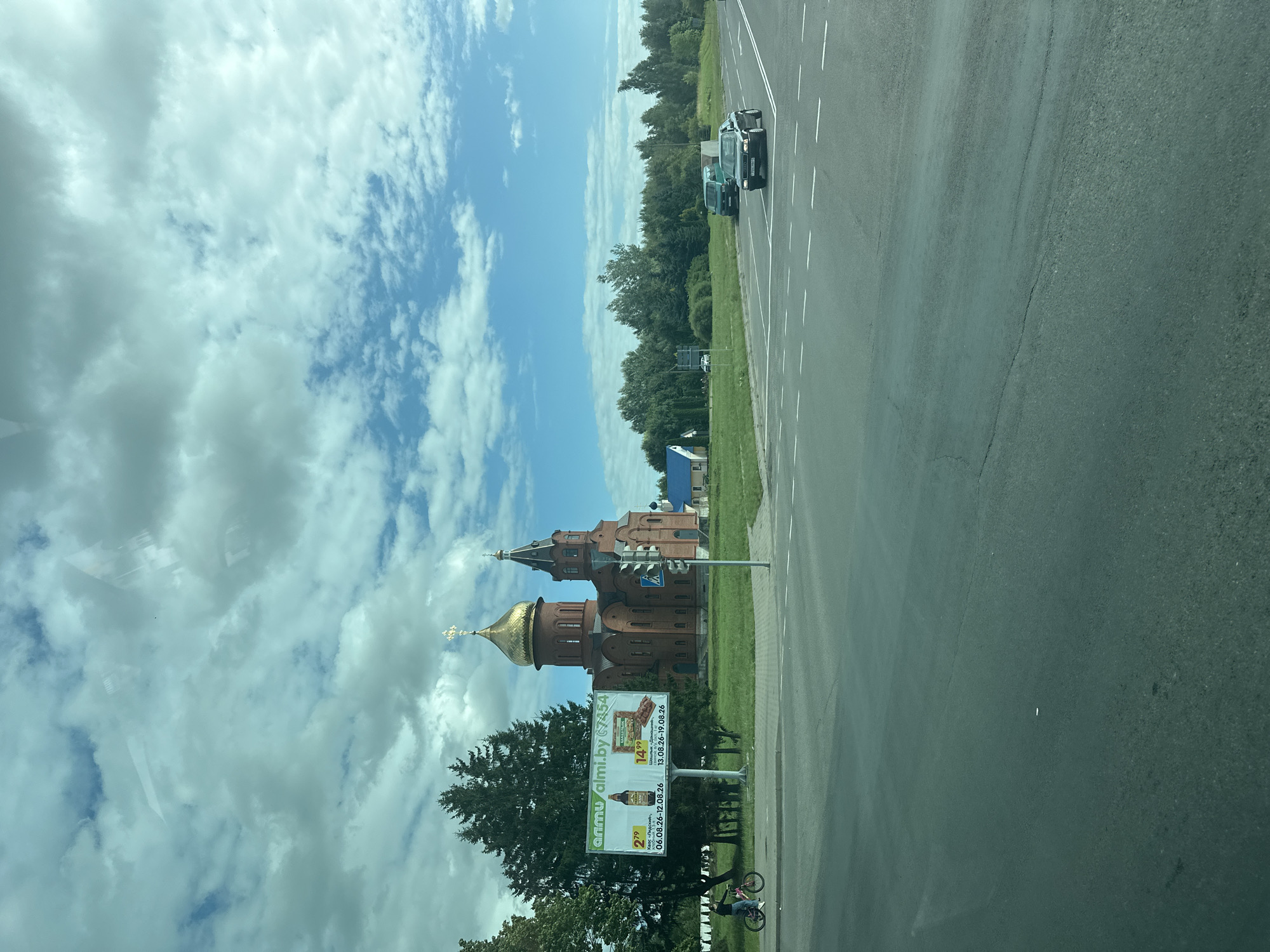 The image shows a red brick Orthodox church with golden domes, located in Maladzyechna, Belarus. A billboard for "almi.by" is visible to the left of the church, displaying prices for beer and snacks. A person on a bicycle is riding on a sidewalk in front of the billboard. The church is situated on a grassy area with trees in the background. A road with white lane markings is in the foreground, with cars driving on it. The sky is blue with scattered white clouds. The overall scene depicts a semi-urban environment with a prominent religious building and everyday street activity.