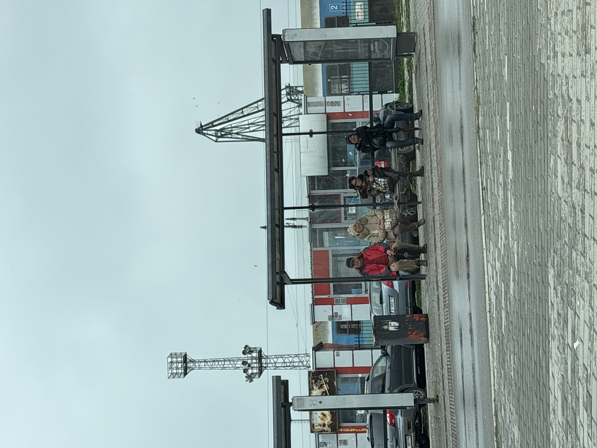 The photo depicts a bus stop in Varna, Bulgaria. Four people are seated on the bench within the shelter, facing right. The person closest to the camera is wearing a red jacket, the next a beige coat. The other two are wearing darker clothing.
Behind the bus stop, there are parked cars. The background features a building with a mixture of red, white, and blue panels. The sky is overcast and grey. Metal structures, likely part of the city's infrastructure, are visible rising into the sky. The pavement is wet and appears to be constructed from light-colored rectangular stones.
