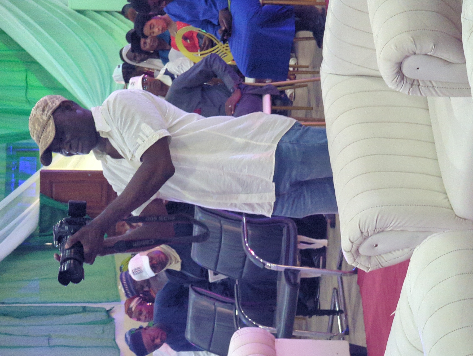 In Ungwan Liman, Nigeria, a man wearing a tan cap, a white short-sleeved shirt, and blue jeans is holding a camera, possibly taking a photograph. The environment appears to be indoors, with green drapes visible on the left. Several other people are seated in the background, including a person in a blue garment with a yellow, hoop-shaped object. Stacks of white, rolled-up materials are on the right, while office-style chairs are visible in the middle ground, occupied by people who may be observing the photographer.