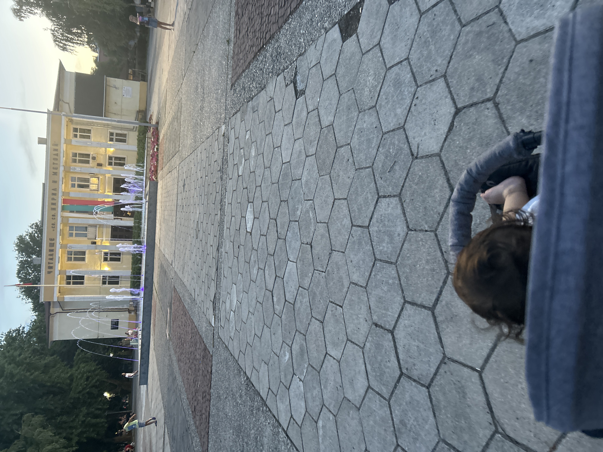 In the city of Parvomay, Bulgaria, on a hexagonal stone-paved plaza, a fountain with colored lights is operational in front of a yellow building. The building has a flag pole with a Bulgarian flag at its apex and text above its entrance. Several children and adults are visible in the background, some walking and others seated. The foreground shows a child's head and feet in a stroller, positioned to capture the scene. The lighting suggests it is either early morning or late evening.