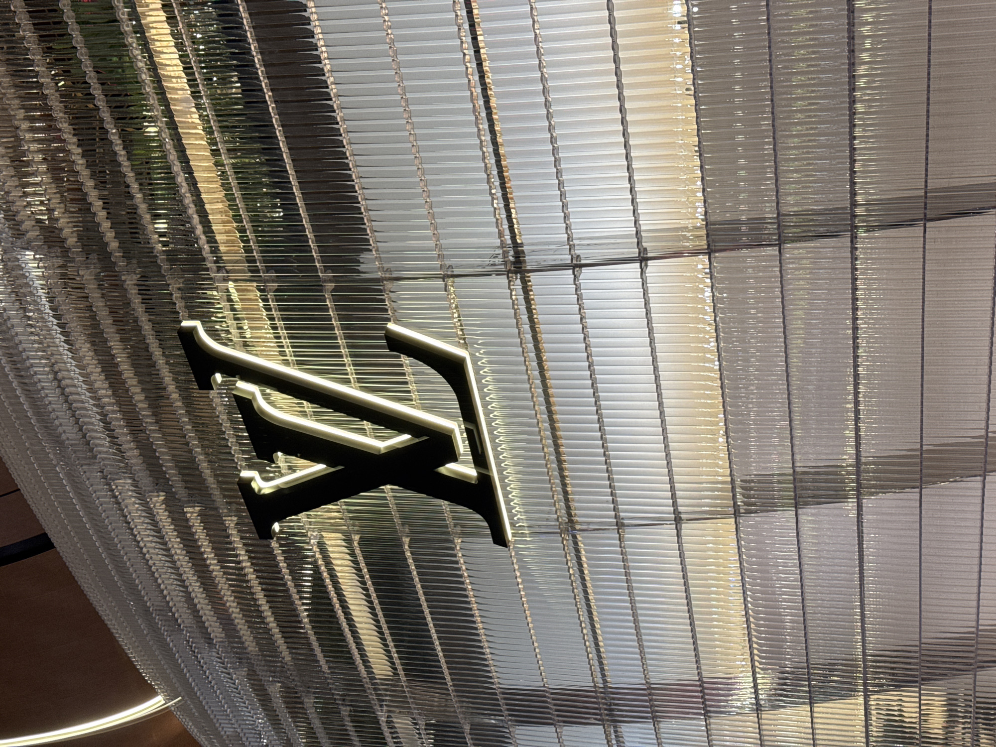The image shows a close-up of the Louis Vuitton storefront in Doha, Qatar. The facade of the building is covered in a translucent, ribbed material that creates a textured effect. A large, illuminated Louis Vuitton logo is prominently displayed on the facade. The lighting suggests it is nighttime or dusk, with interior lights glowing from behind the facade and illuminating the logo. The overall impression is one of modern luxury and sophisticated branding.