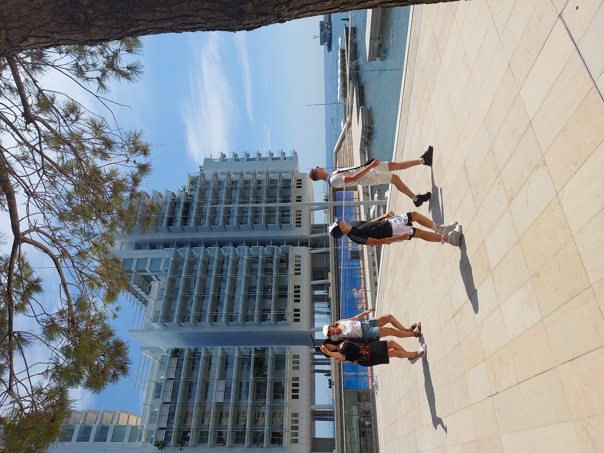 The image depicts a sunny day with a clear blue sky in Monaco. In the foreground, a paved walkway is occupied by five people. On the left, two women are walking side-by-side. The woman on the left is wearing a black dress and a dark hat, holding a smartphone up as if taking a selfie. The woman next to her is wearing a white top and denim shorts, with a white cap on her head. To their right, a young boy in a black t-shirt and white shorts with a black and white cap is walking. Further to the right, a man in a white t-shirt and white shorts is walking, carrying a dark bag over his shoulder. Behind the people, modern, multi-story buildings with glass balconies are visible. The buildings appear to be residential or hotel complexes. To the right of the buildings, a body of water, likely the Mediterranean Sea, is visible. There are some boats and structures in the water, and white umbrellas are set up on a platform near the water's edge. The scene conveys a leisurely atmosphere, suggesting a popular tourist or residential area in Monaco.