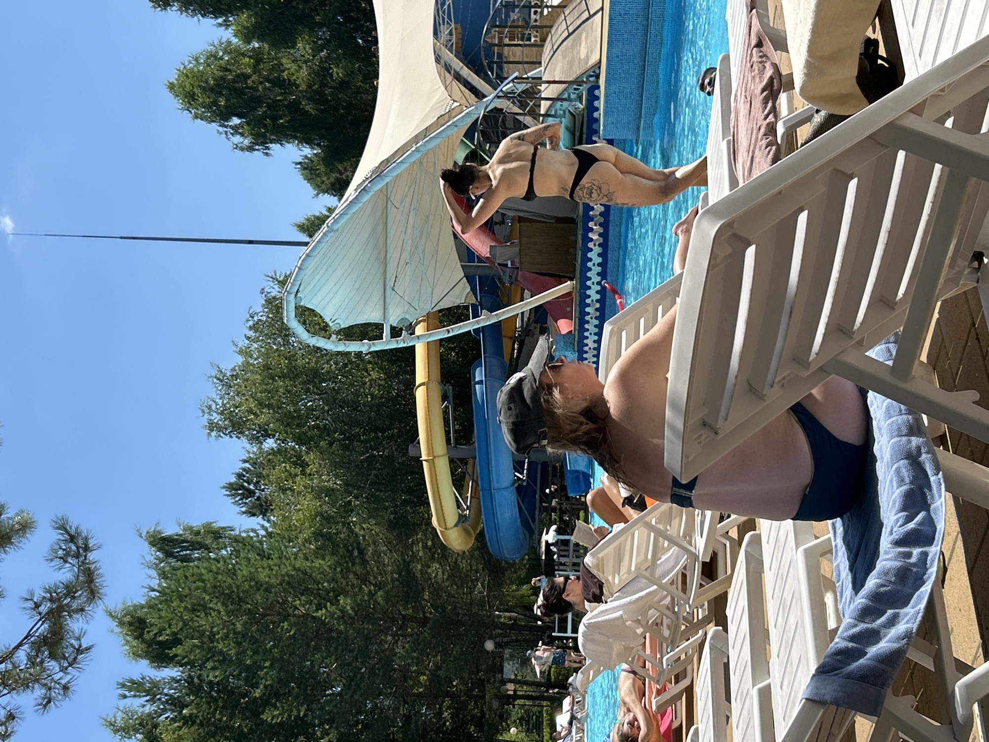The image depicts a sunny day at a water park in Togliatti, Russia. In the foreground, several white lounge chairs are arranged around a blue swimming pool. A woman wearing a dark swimsuit and a baseball cap is seated on one of the chairs, facing away from the camera. Another person is partially visible on a lounge chair to the left. In the mid-ground, a woman in a black bikini with tattoos is standing near the edge of the pool, with her back to the camera, adjusting her hair. Behind her, a large blue and yellow water slide is visible. Trees surround the water park. The sky is clear and blue.