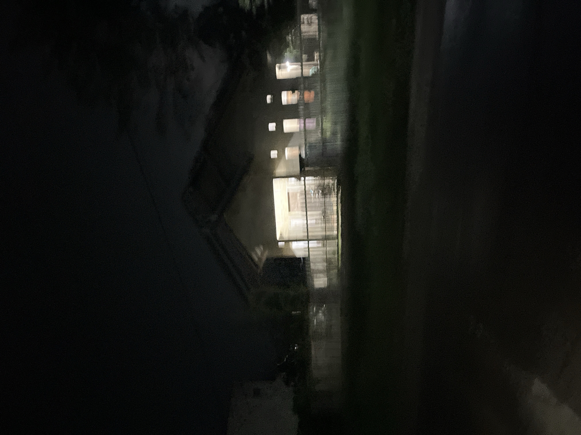 The image shows a house at night. The house is light brown with a dark roof. Several windows are illuminated, suggesting occupancy. There is a fence in front of the house, and trees are visible in the background. The foreground is dark and appears to be a grassy area. The image is grainy due to low light conditions. The location is identified as Landuh, Indonesia.