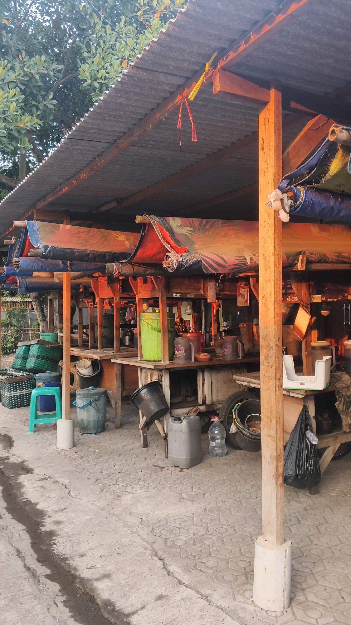 This image depicts an outdoor market stall with a corrugated metal roof supported by wooden beams. The stall is set up with wooden counters displaying various containers, including a large green bucket, several plastic pitchers, and smaller buckets. In front of the stall, there are several large plastic containers, a blue plastic stool, and woven baskets. A black trash bag is visible to the right. The ground is paved with hexagonal concrete tiles, and the area appears to be in an outdoor setting, possibly a market in the city of Margadadi, Indonesia, given the provided context. The lighting suggests it might be late afternoon or early morning, with warm light hitting the wooden structures.